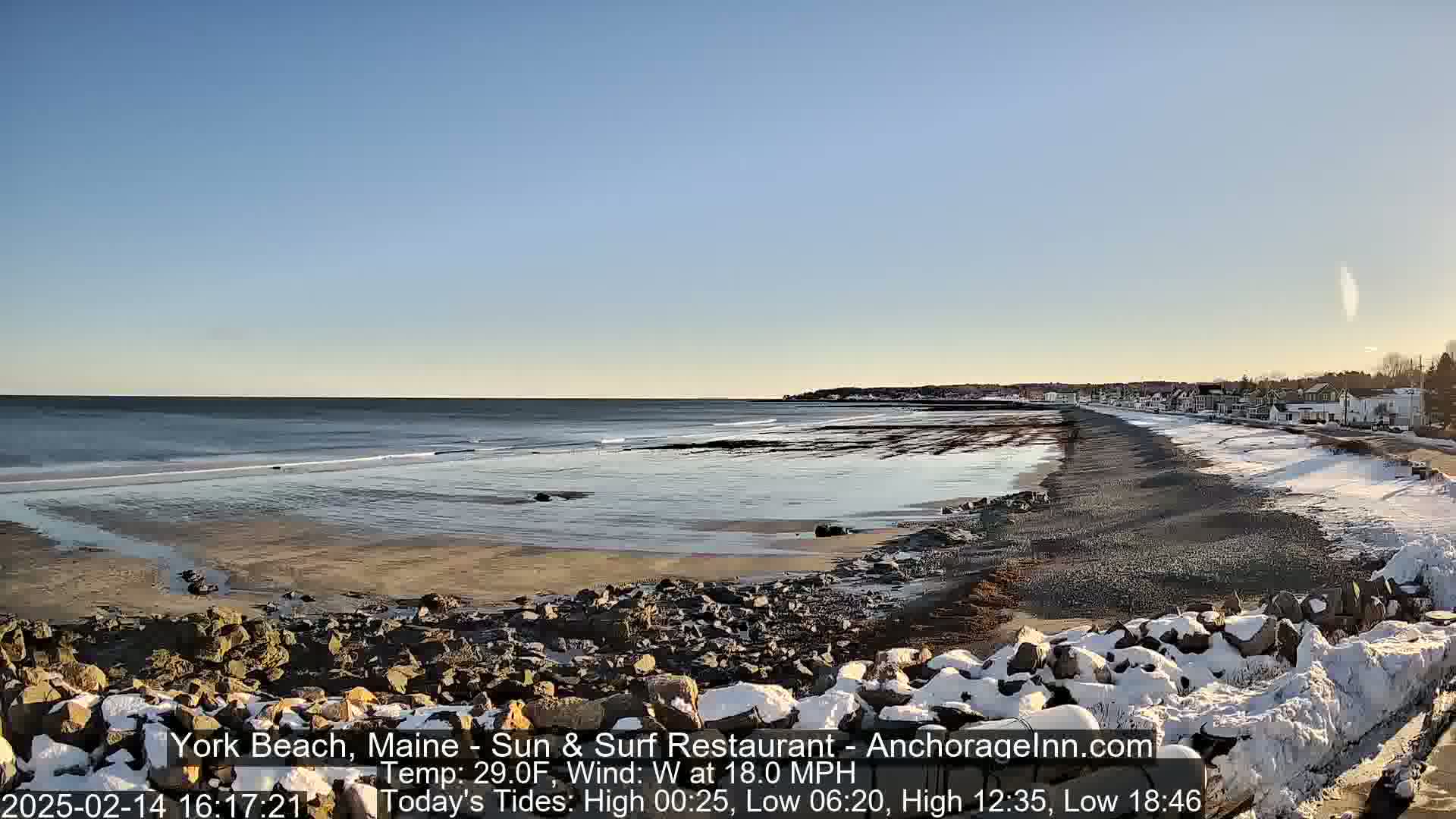 A partly snowy beach with a rocky shoreline and calm ocean under a clear, bright sky.