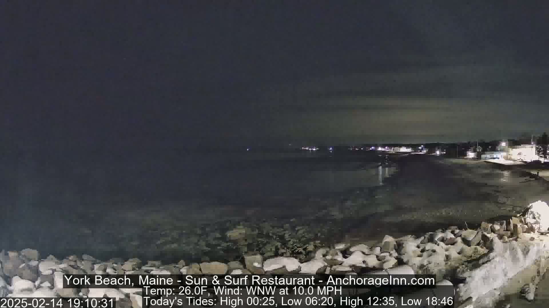 A nighttime view of a dark, rocky beach with a calm ocean and distant, dimly lit buildings under a clear sky.