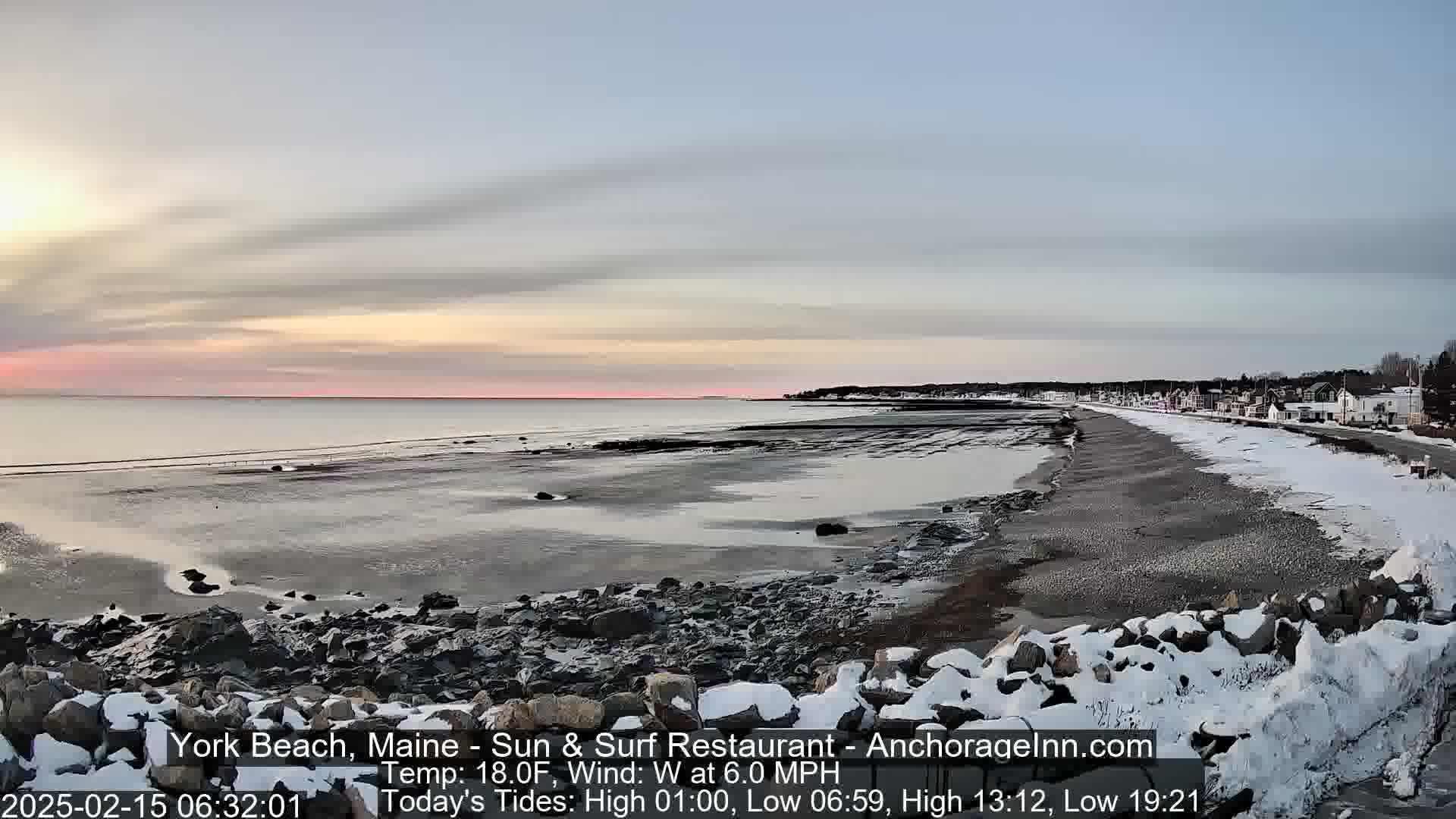 A partially snow-covered rocky shoreline stretches alongside a calm, partially exposed sandy beach at sunrise under a pale sky.