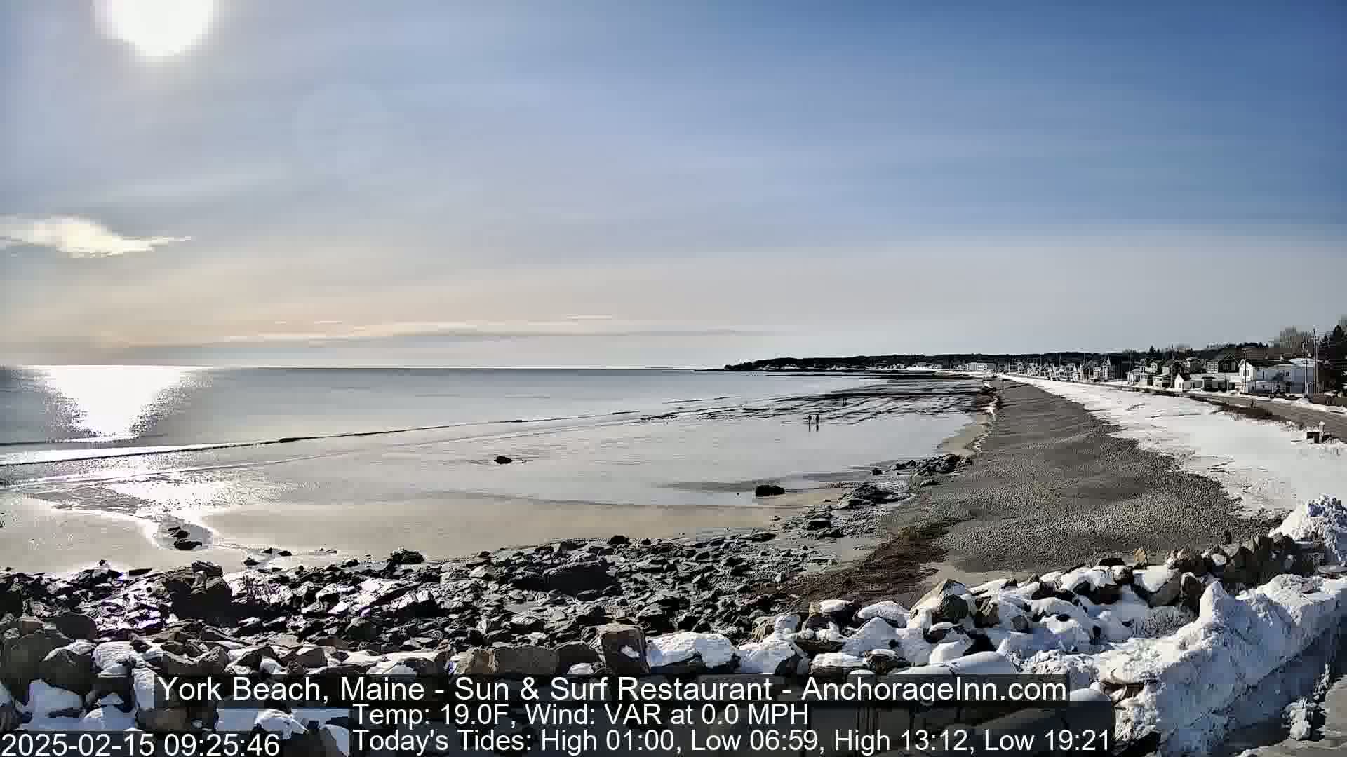 A mostly sunny day reveals a partially frozen, snow-dusted beach with a calm ocean and a small town in the background.
