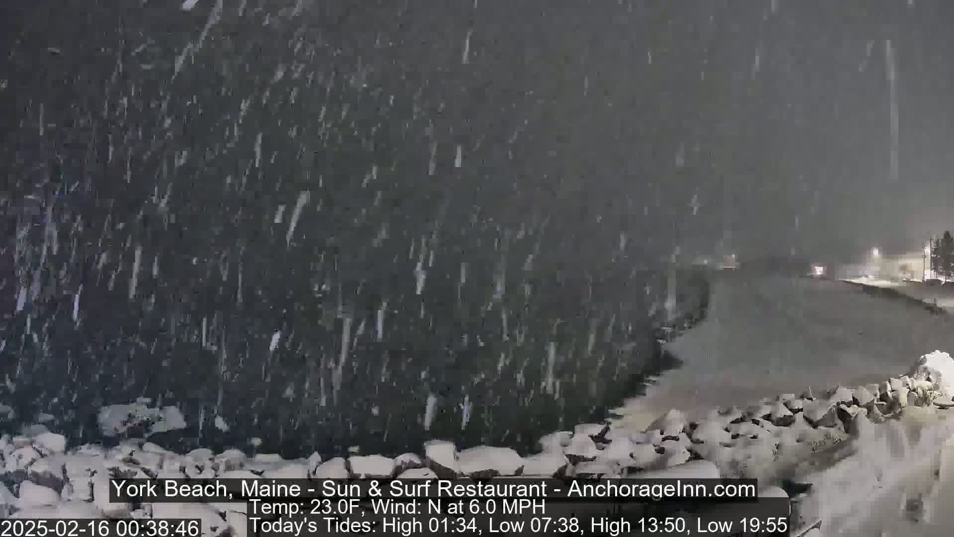 A nighttime scene of heavy snowfall over a rocky shoreline next to a road.