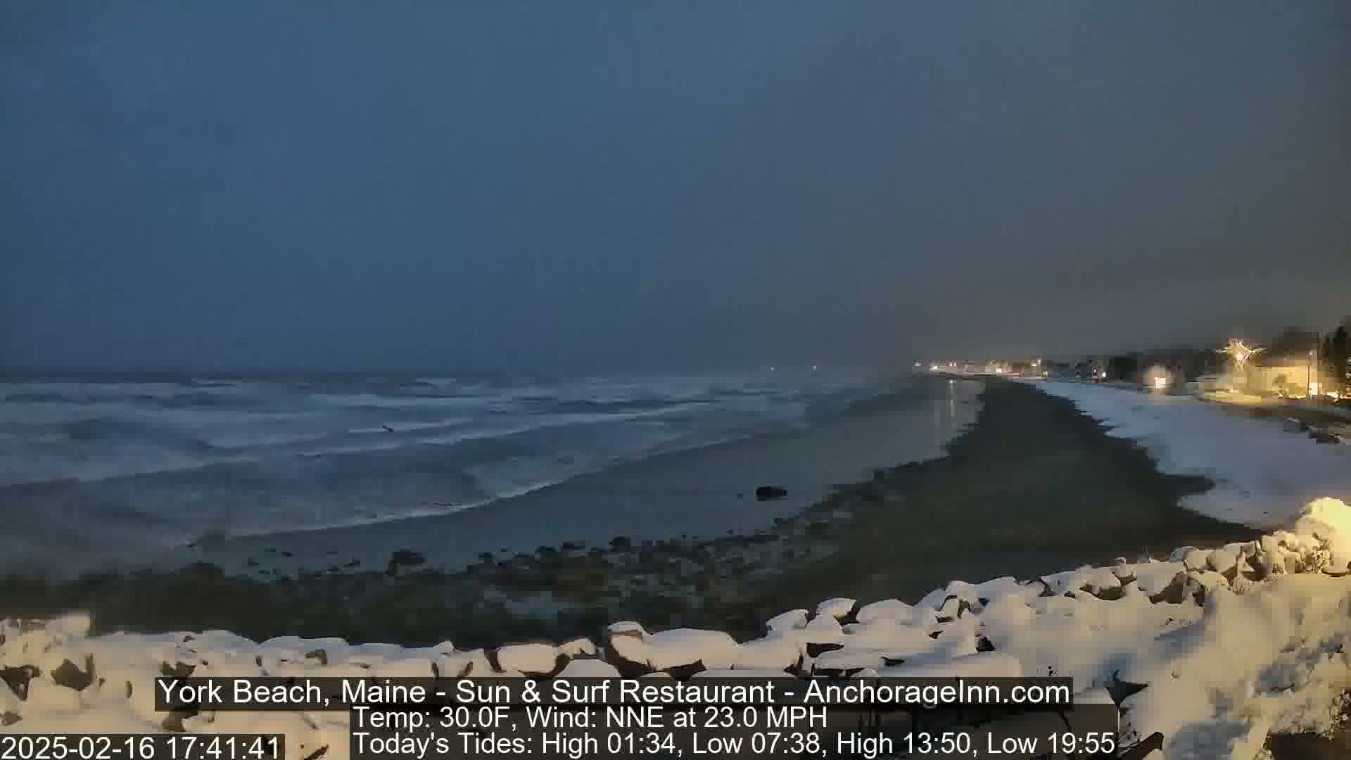 A snow-covered beach at night with dark, choppy waves under a gray sky.