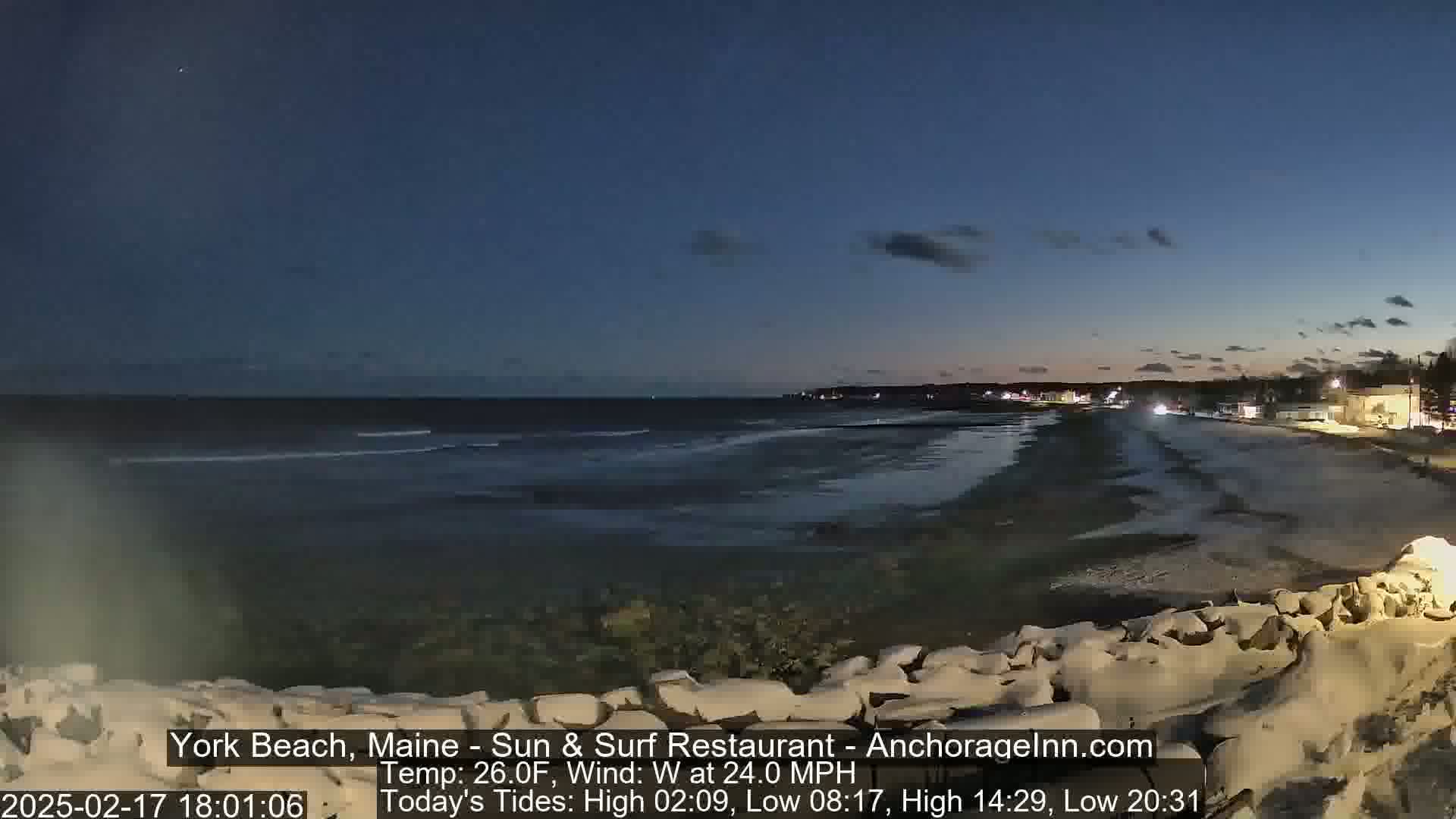 A dark, windy evening at the beach shows a mostly calm ocean with a few small waves lapping at the shore, under a twilight sky.