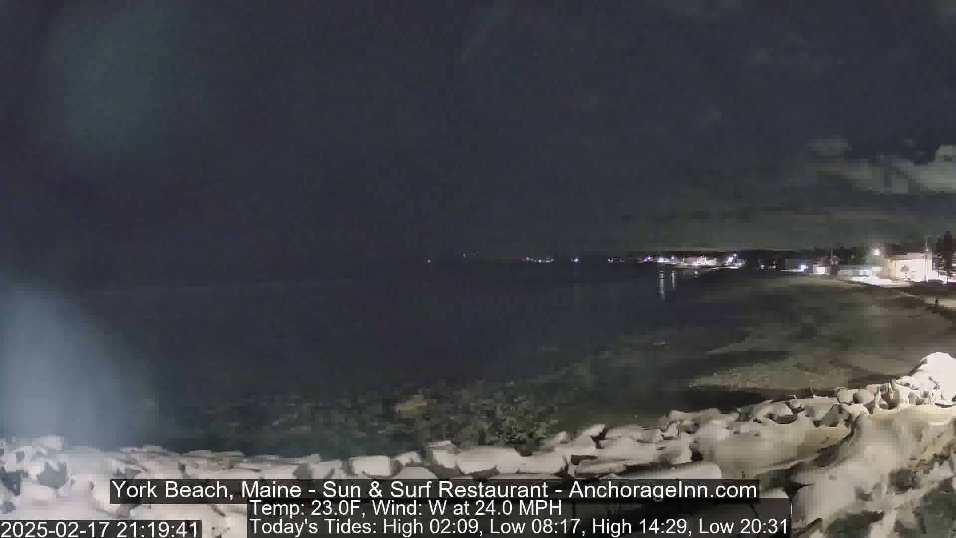 A nighttime view of a dark, rocky shoreline and a calm, dimly lit ocean under a cloudy sky with a 24 mph west wind and a temperature of 23°F.