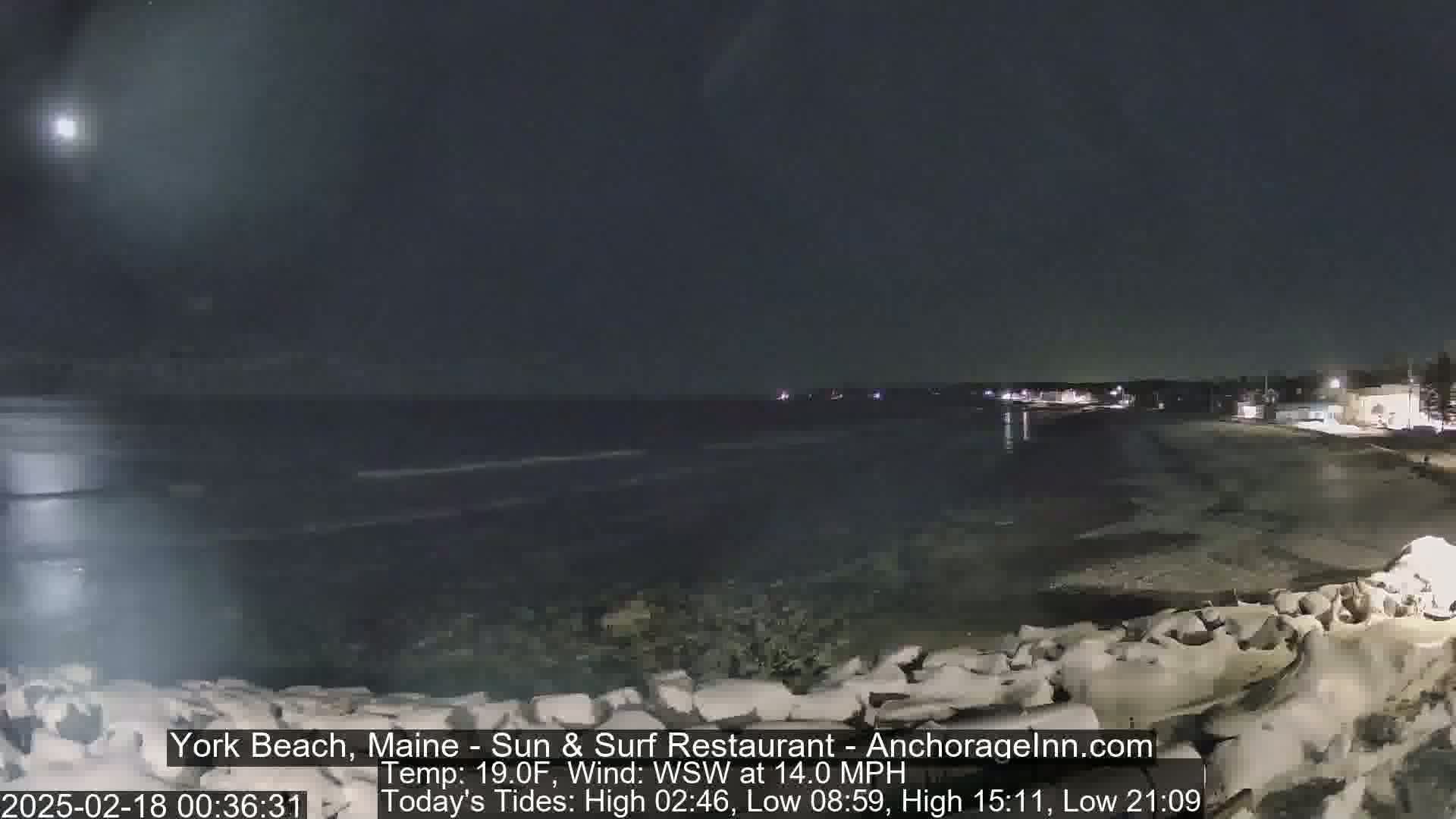 A nighttime view of a dark, wavy ocean shore with a partially visible town in the distance under a mostly clear sky.