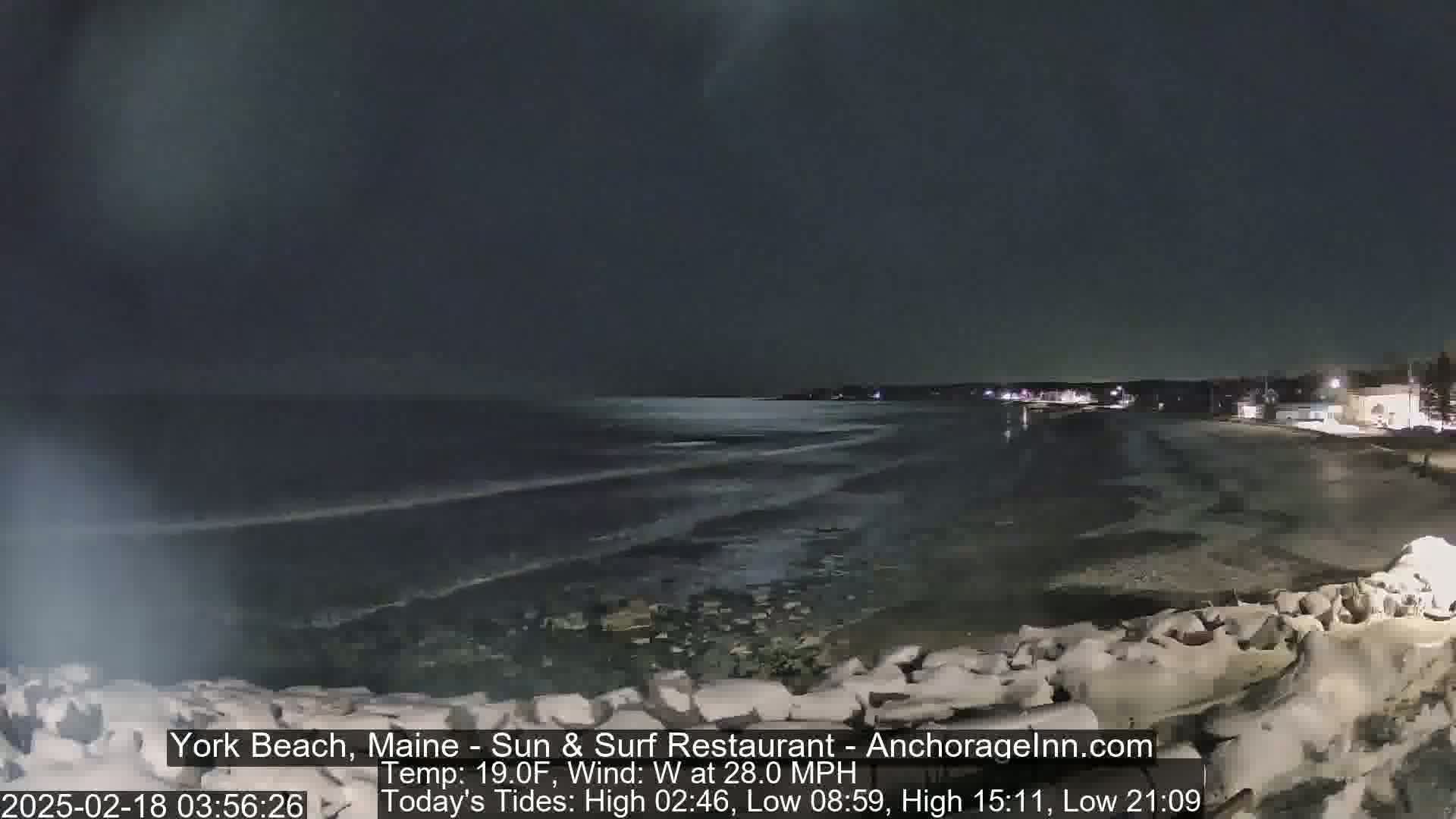 A dark, nighttime beach scene shows a wavy ocean under a mostly clear sky, with a rocky shoreline and distant lights visible.