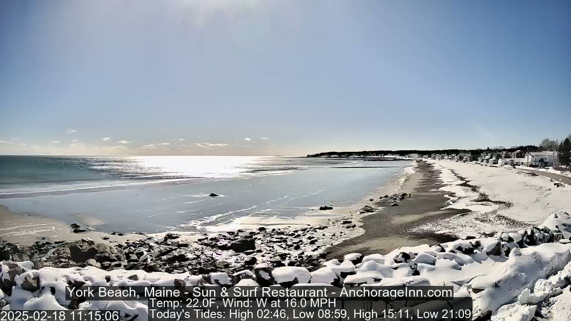 A partially snow-covered beach with a calm ocean under a sunny sky.