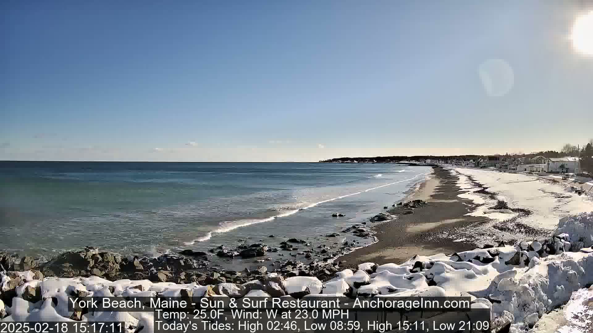 A snow-covered beach with a dark-sand shoreline and a partly-frozen ocean under a clear, sunny sky.