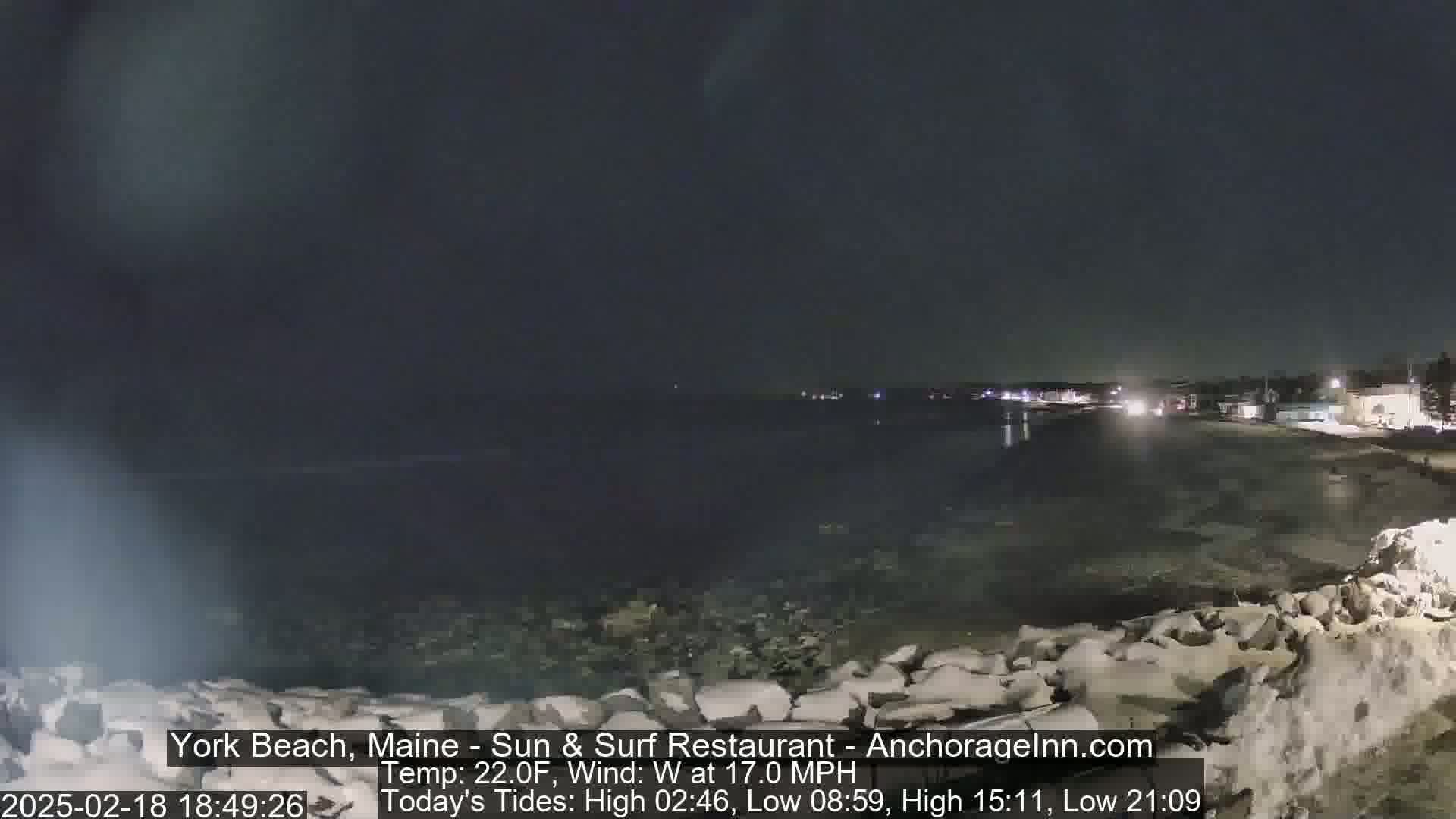 A dark, nighttime beach scene with a calm body of water, a rocky shoreline partially covered in snow, and distant lights on the land, under a mostly clear sky with a 17 mph westerly wind.