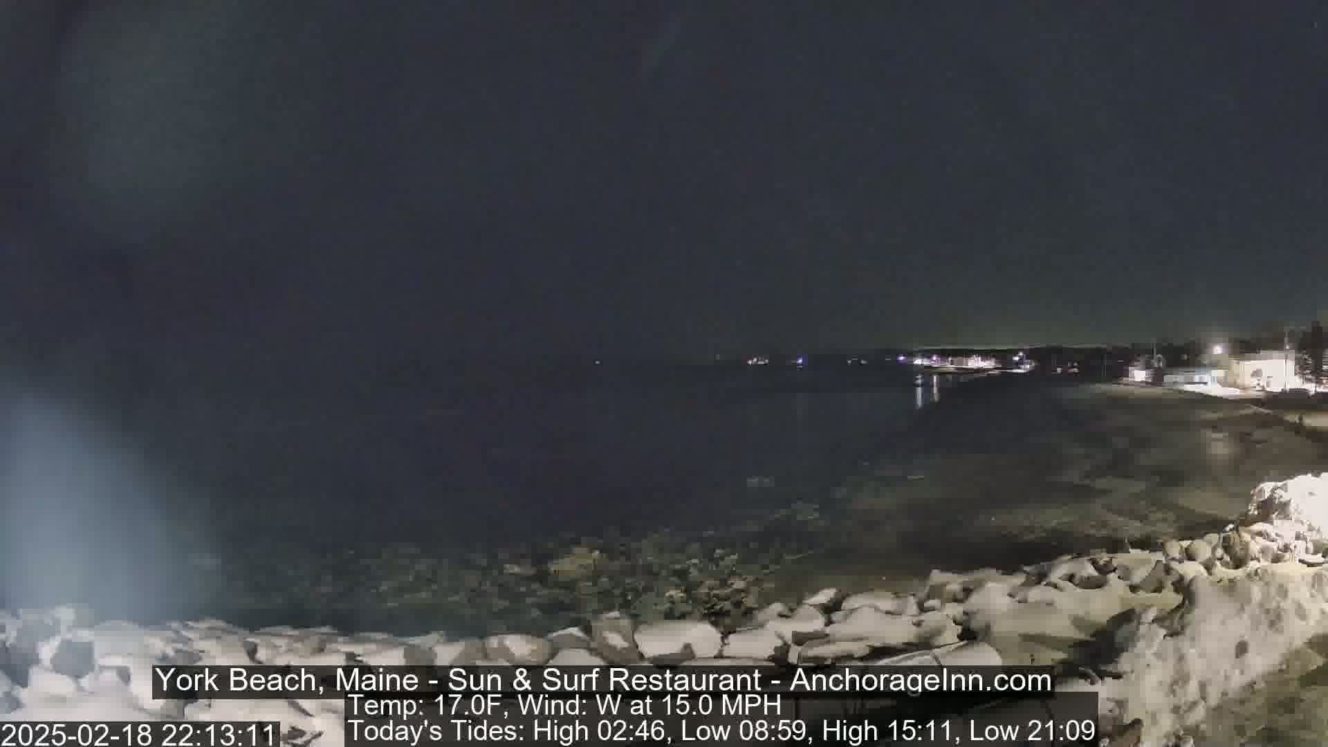 A nighttime view of a calm, dark ocean with a shoreline featuring a rocky barrier, under a clear, dark sky.