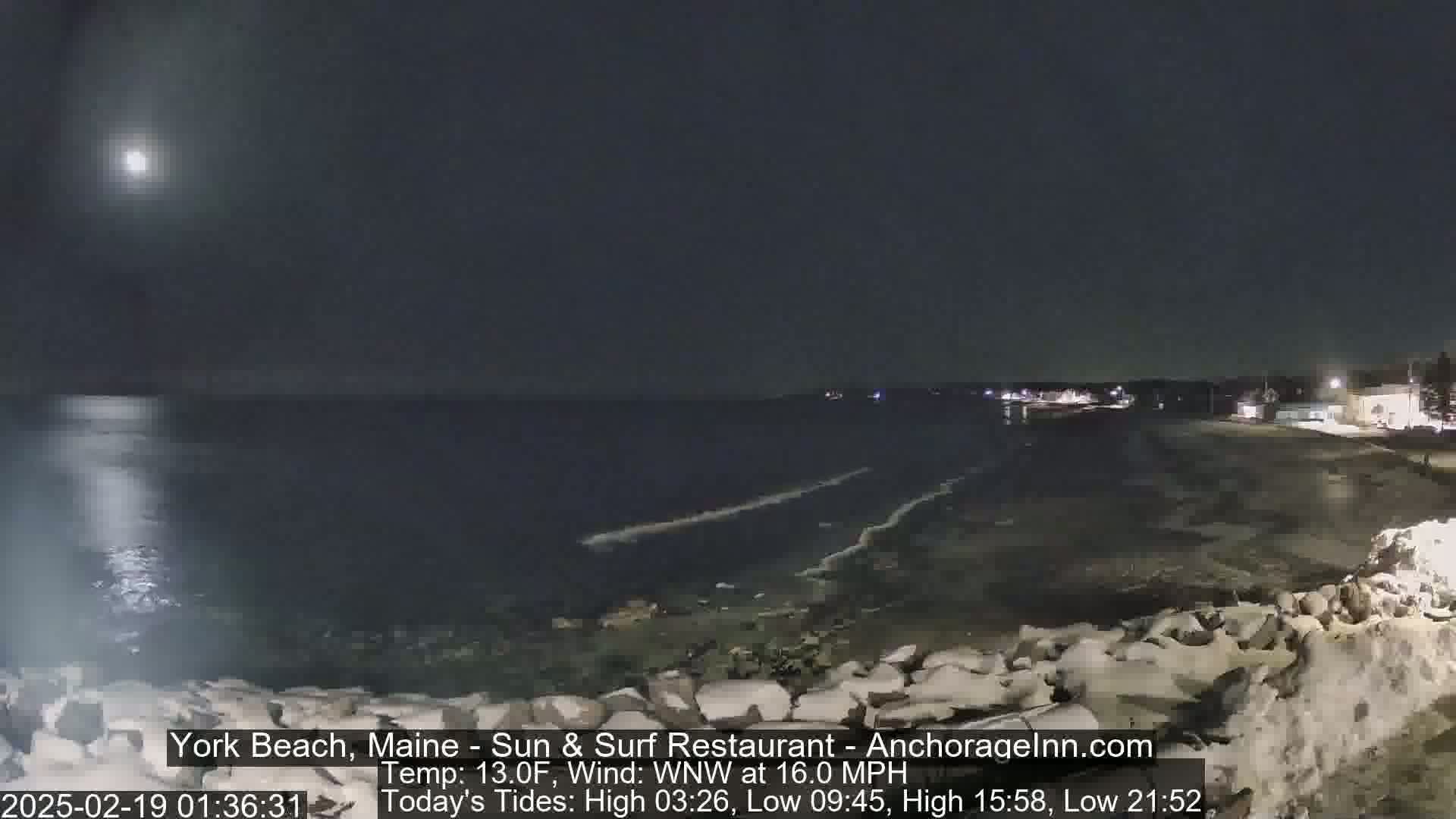 A nighttime coastal scene shows a dark ocean under a bright moon, with a rocky shoreline partially covered in snow and faint lights visible in the distance.
