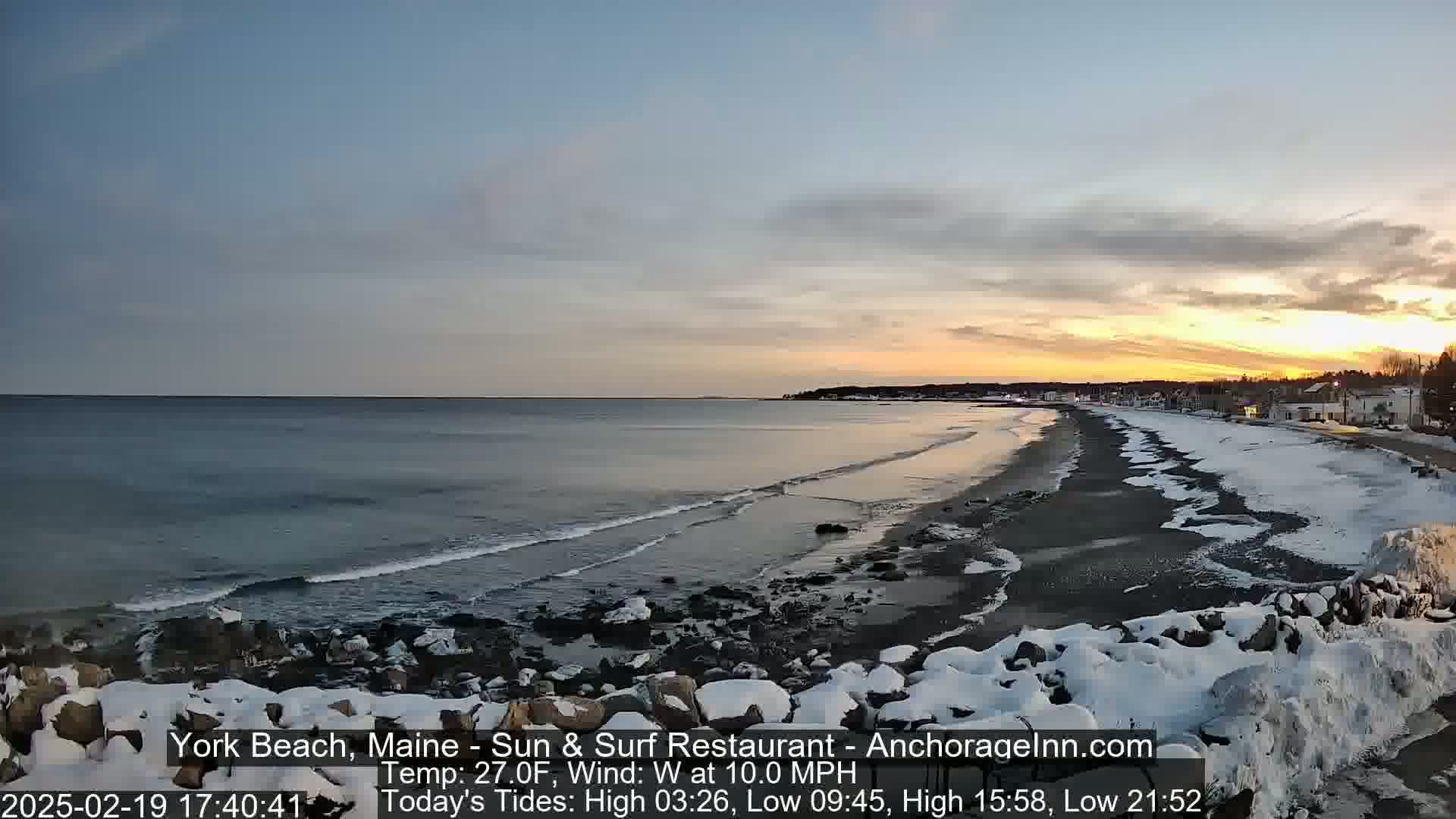 A snowy beach with a calm ocean at sunset under a partly cloudy sky.