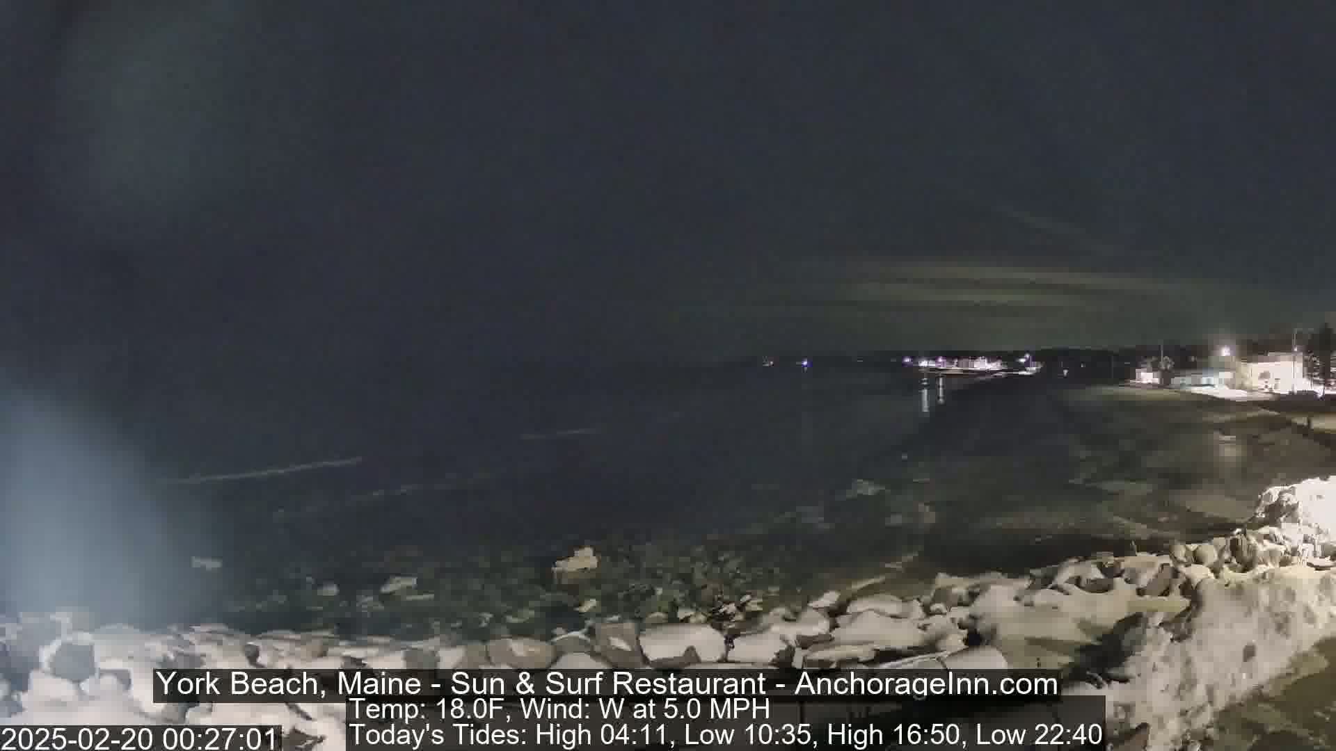 A nighttime view of a snow-covered rocky shoreline with calm dark water and faintly lit buildings in the distance under a dark, cloudy sky.