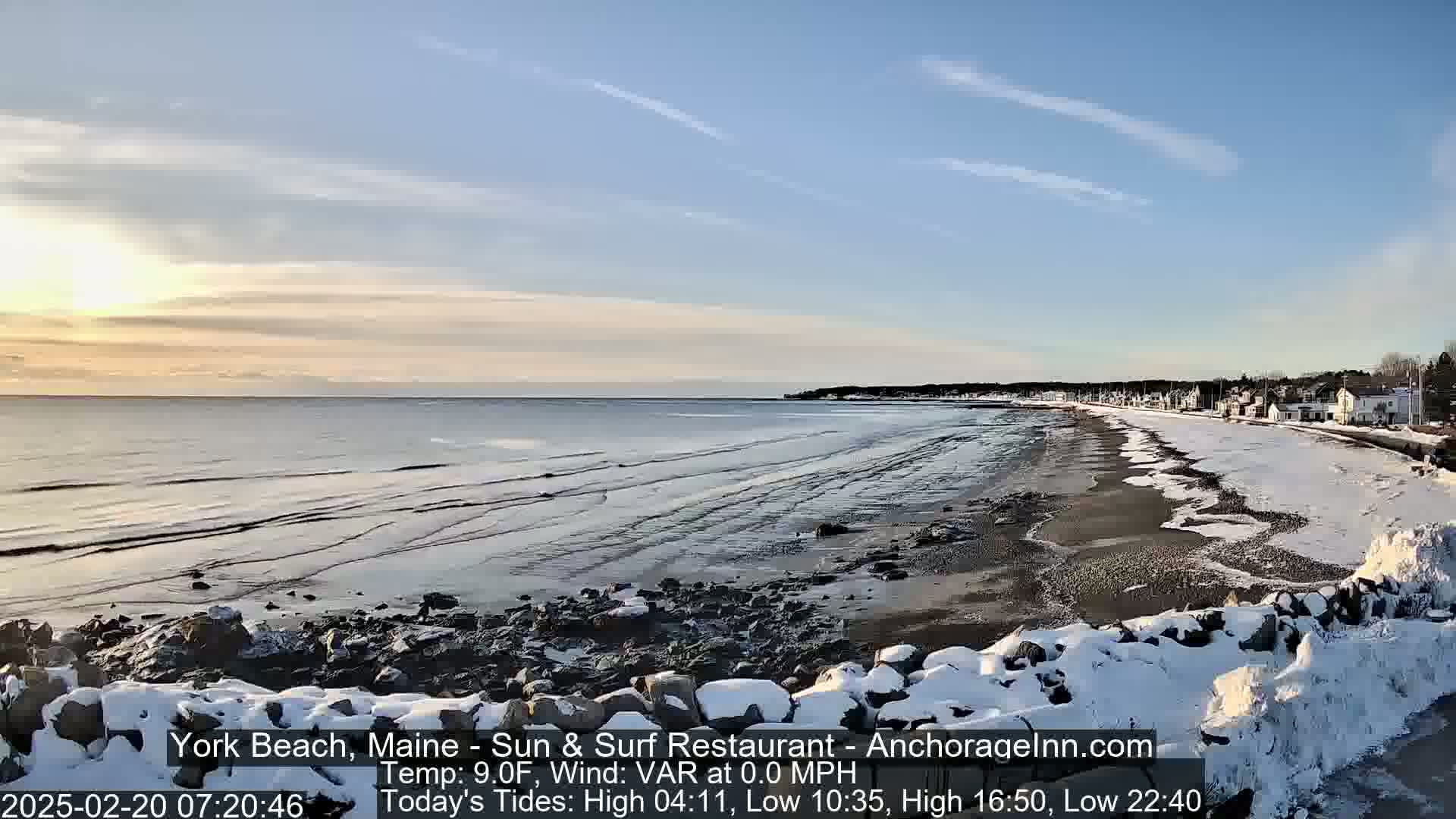 A snow-covered rocky shoreline stretches alongside a calm, sandy beach under a partly cloudy sky at sunrise.