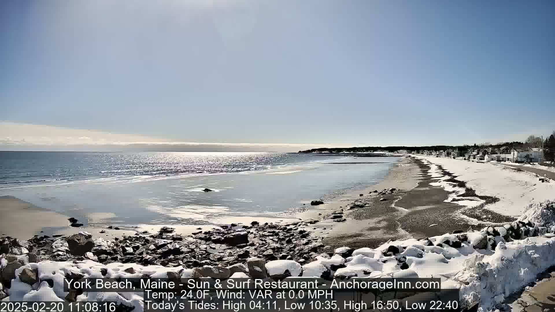 A partially snow-covered beach stretches along a calm, sunlit ocean under a clear blue sky.