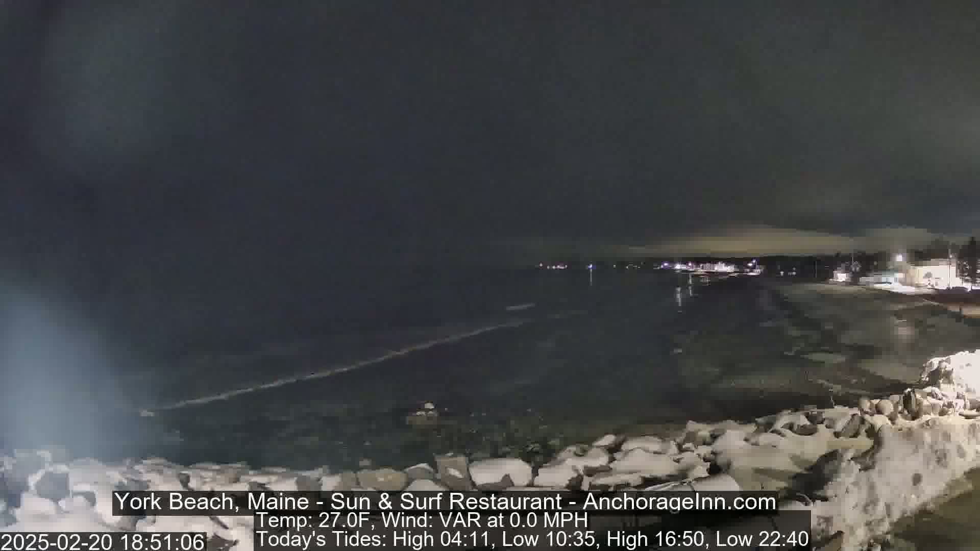 A nighttime view of a dark, calm ocean with a shoreline partially covered in snow and ice under a cloudy sky.