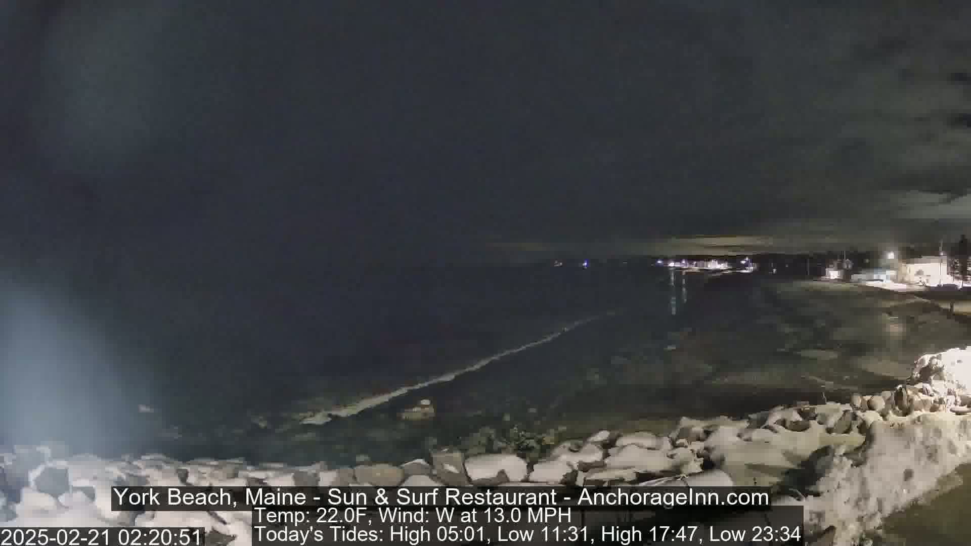 A dark, nighttime coastal scene shows a rocky shoreline partially covered in snow, with a calm, dark body of water extending to a faintly lit town in the distance under a cloudy sky.