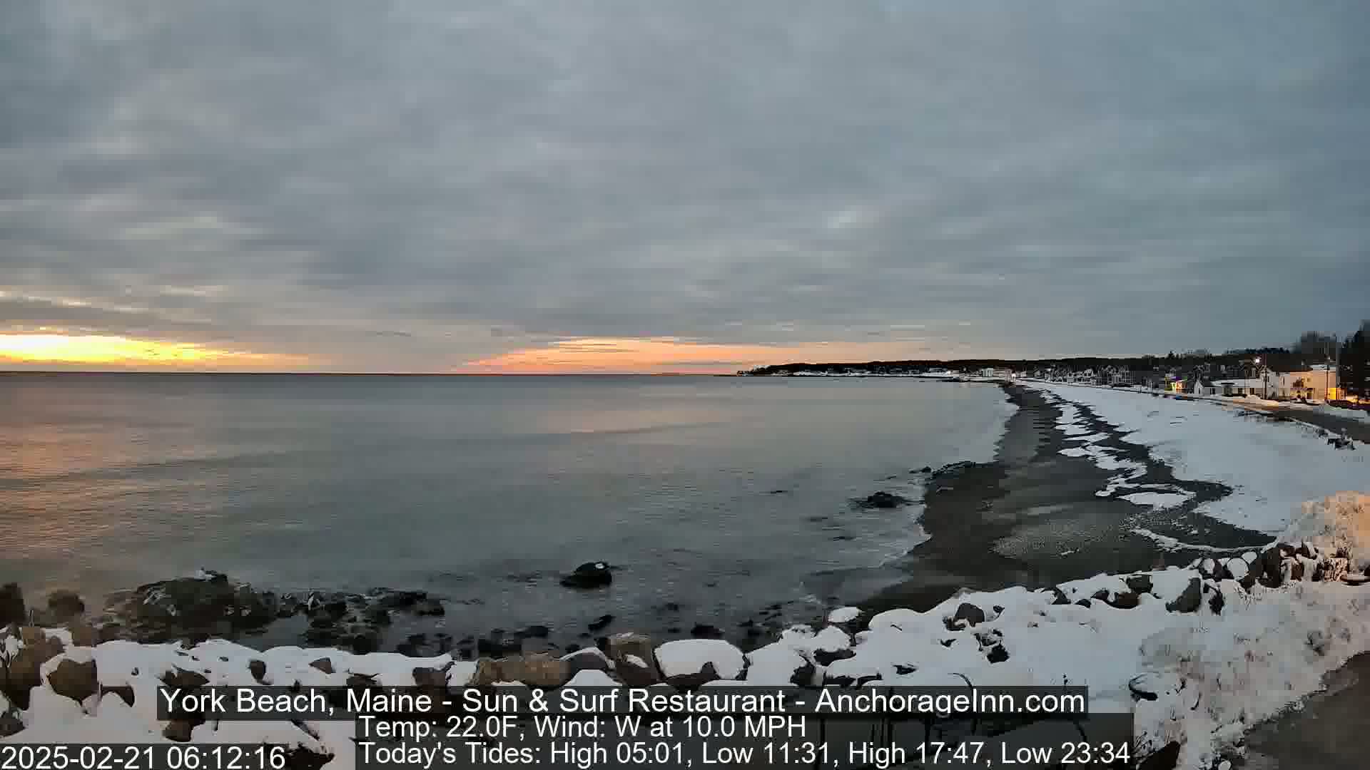 A partly cloudy sunrise over a snow-dusted beach with calm water and a town in the distance.