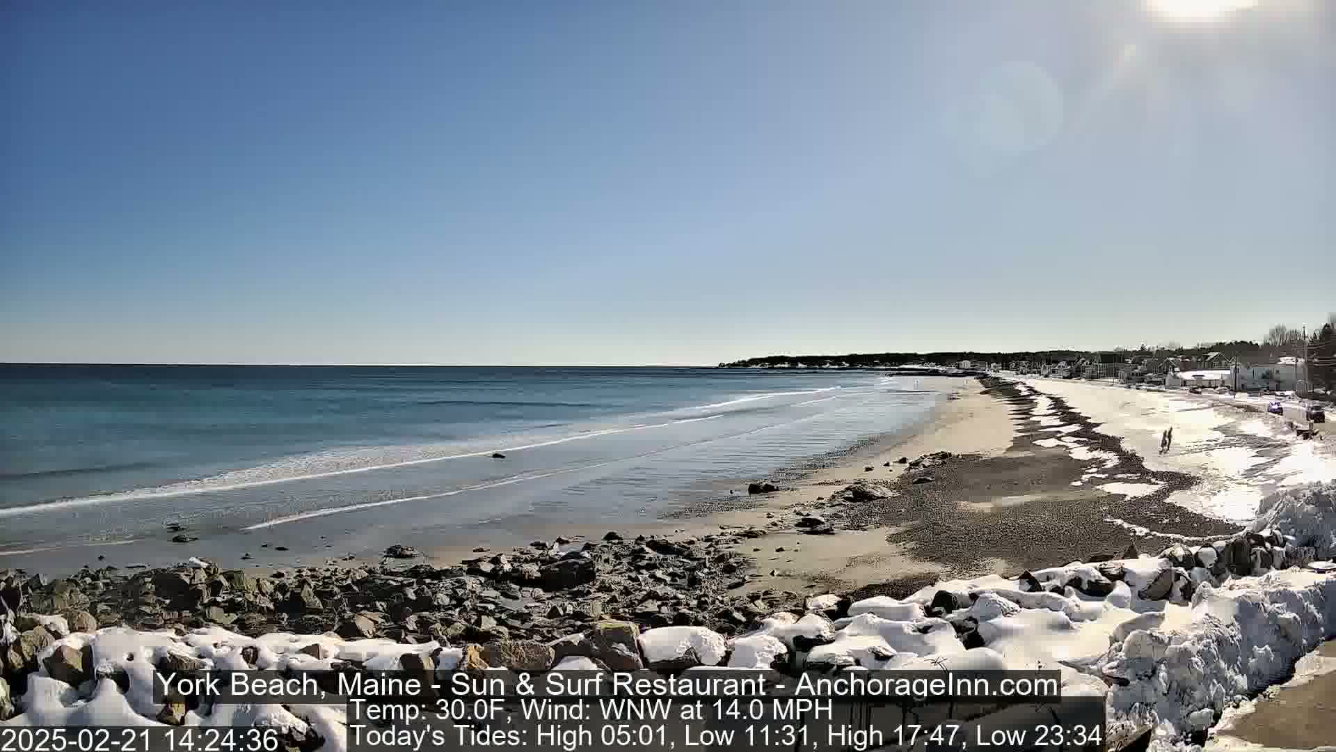 A sandy beach with patches of snow and ice stretches alongside a calm ocean under a clear blue sky.