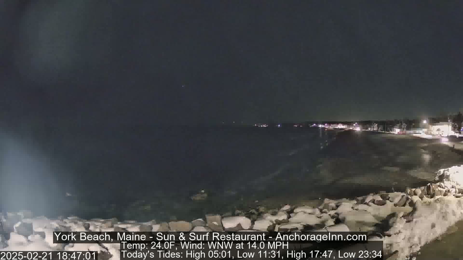 A dark, nighttime beach scene shows a partially frozen, wavy ocean, a snow-covered rocky shoreline, and faintly lit buildings in the distance under a clear sky.