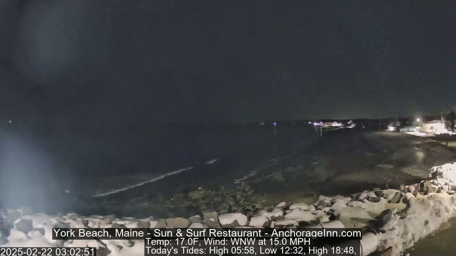 A dark, nighttime beach scene with a partially frozen, rocky shoreline, gently lapping waves, and a calm ocean under a cloudy sky.