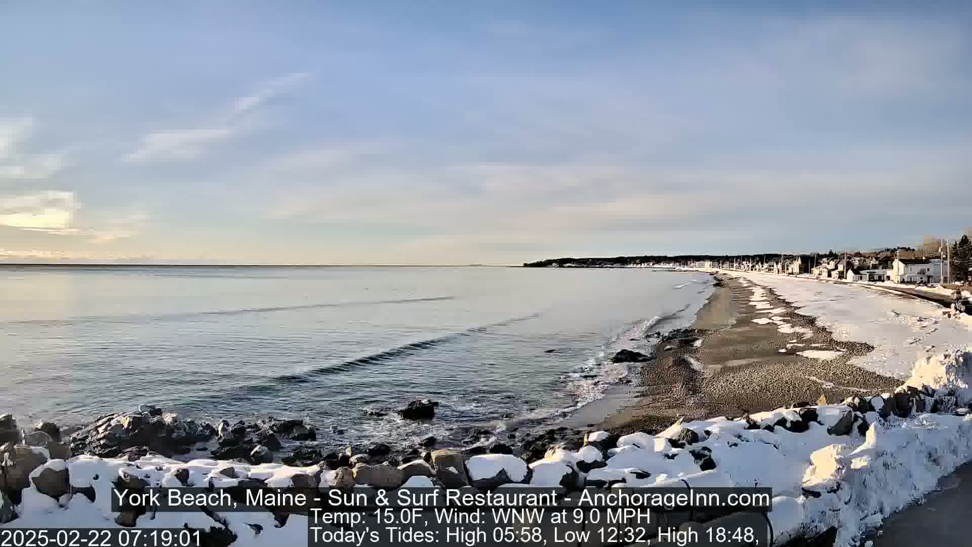 A snow-covered beach stretches along a calm, dark-blue ocean under a mostly clear sky.