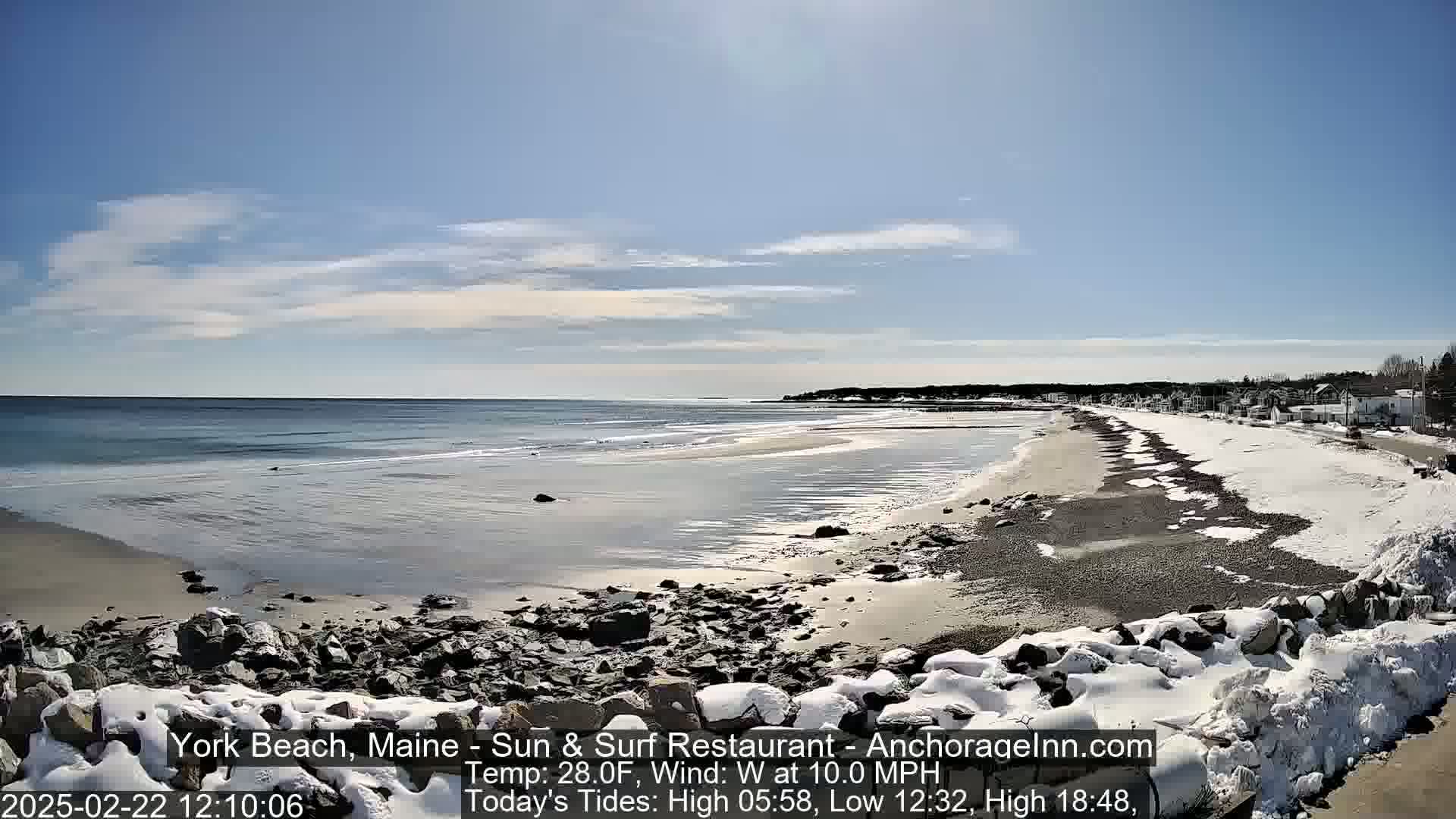 A partly sunny, cold day at a snow-dusted beach with a calm ocean and a town visible in the distance.