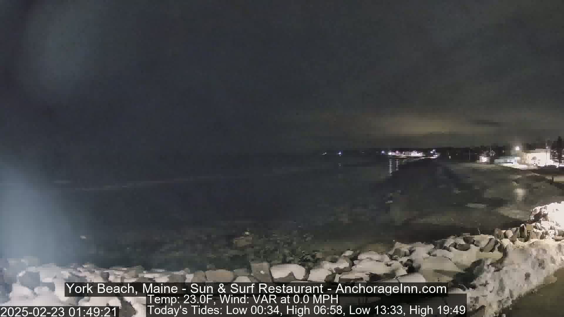 A nighttime view of a snow-covered rocky shoreline and a dark, calm body of water under a clear, dark sky.