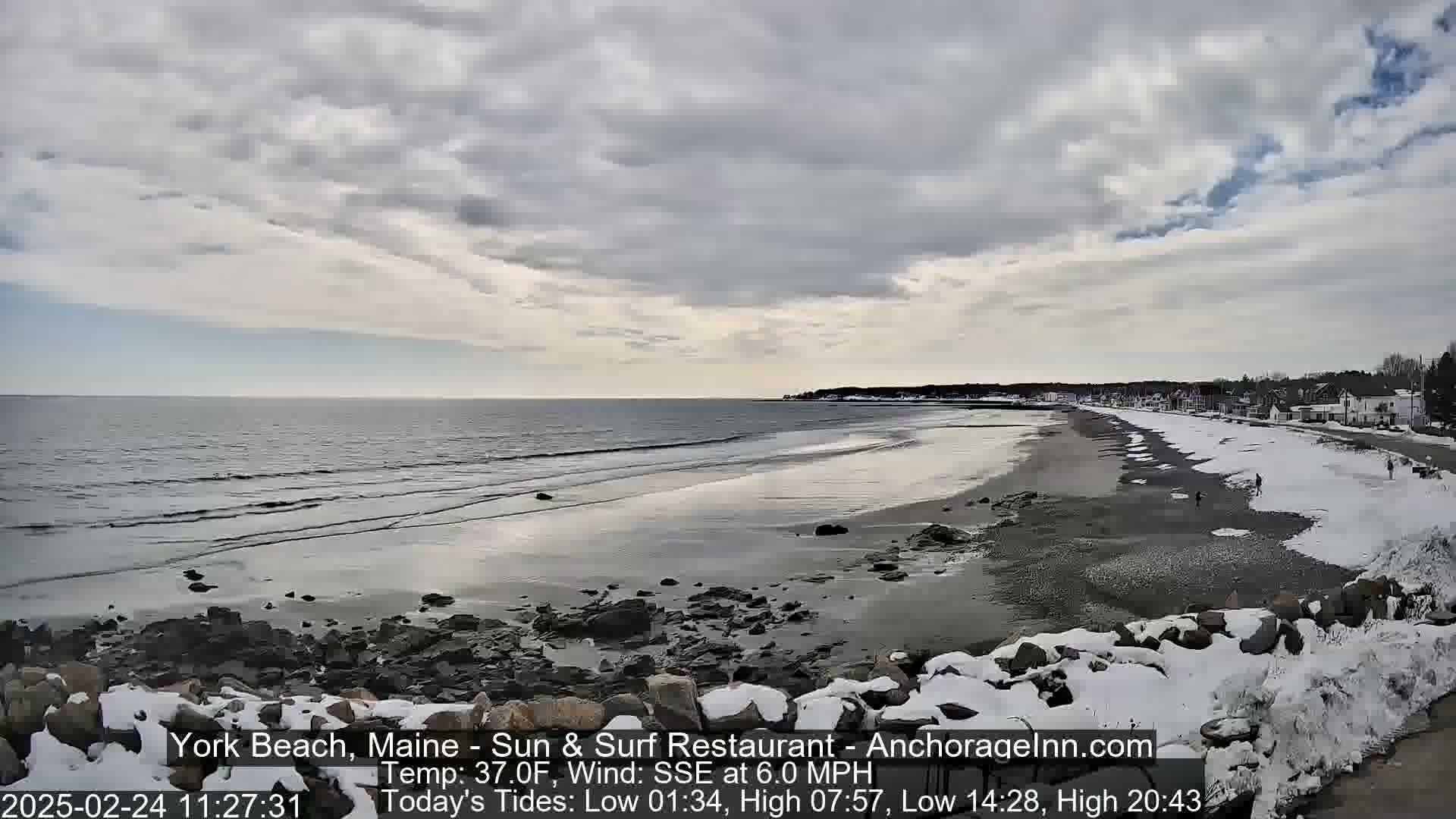 A partially cloudy day at a snow-dusted beach shows a calm ocean with small waves lapping the shore.