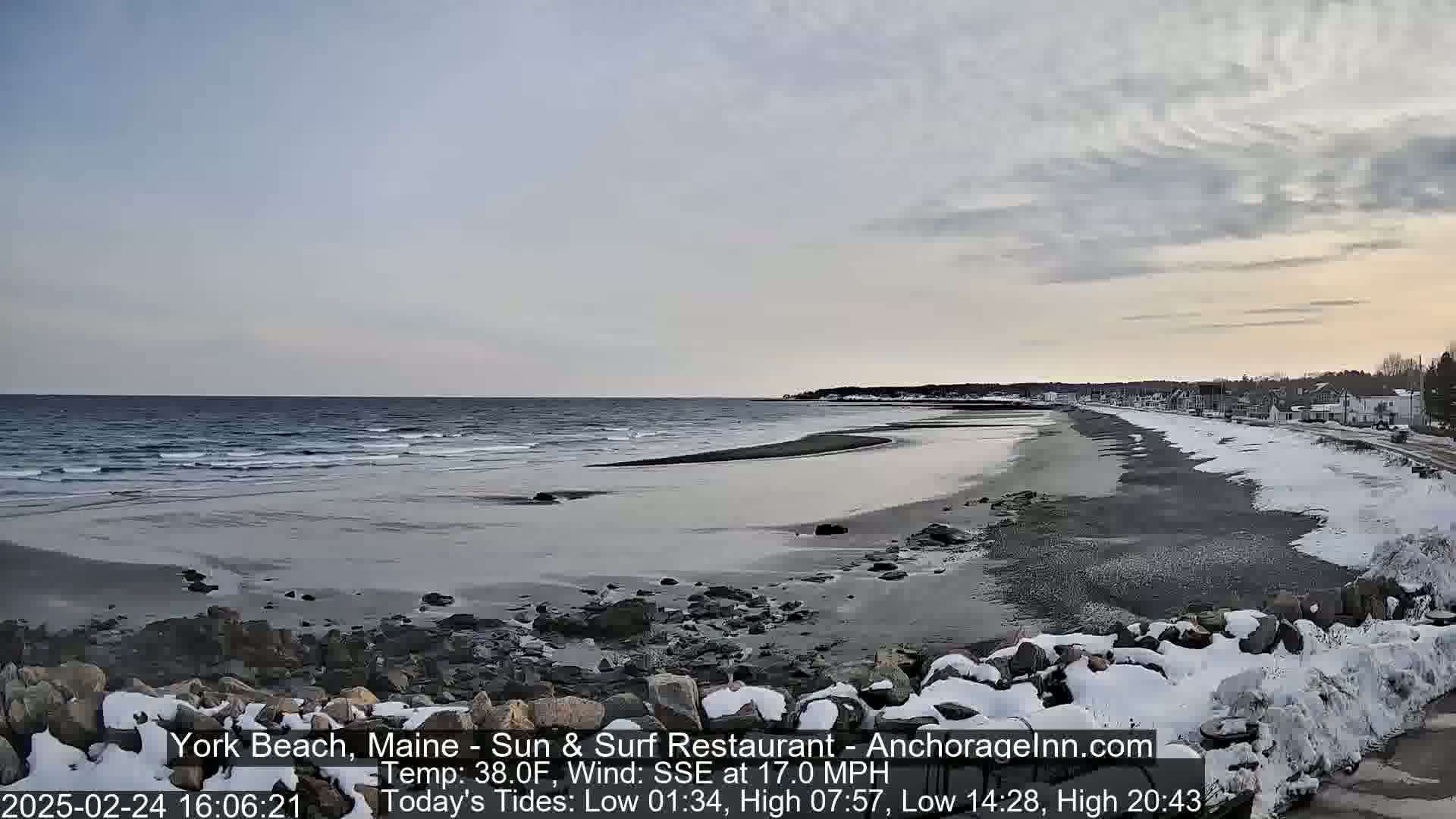 A partially snow-covered beach with a calm ocean under a cloudy sky.