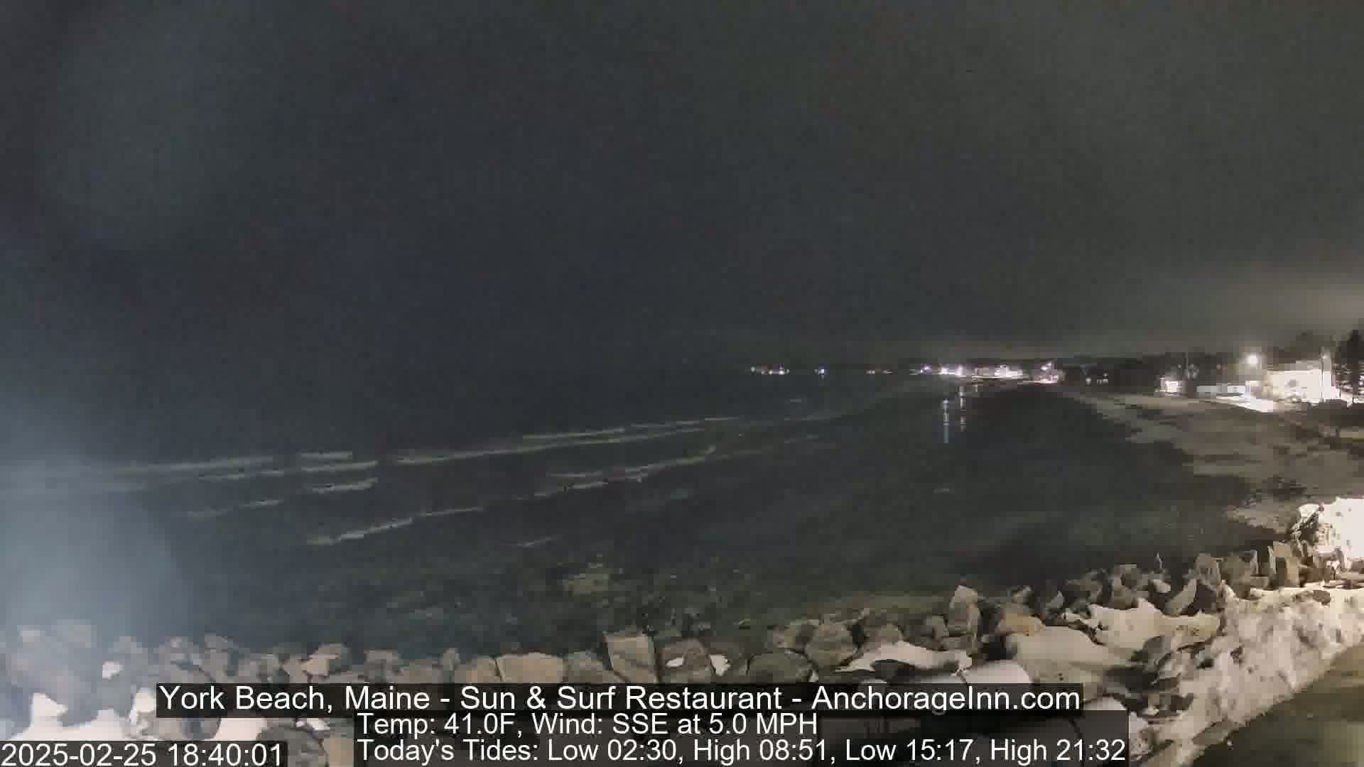 A dark, nighttime view of a beach with gently lapping waves, a rocky shore, and distant lights from buildings along the coast.