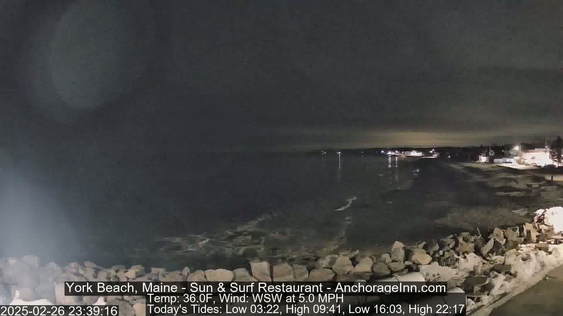 A nighttime view of a dark, calm ocean lapping against a rocky shore under a cloudy sky.