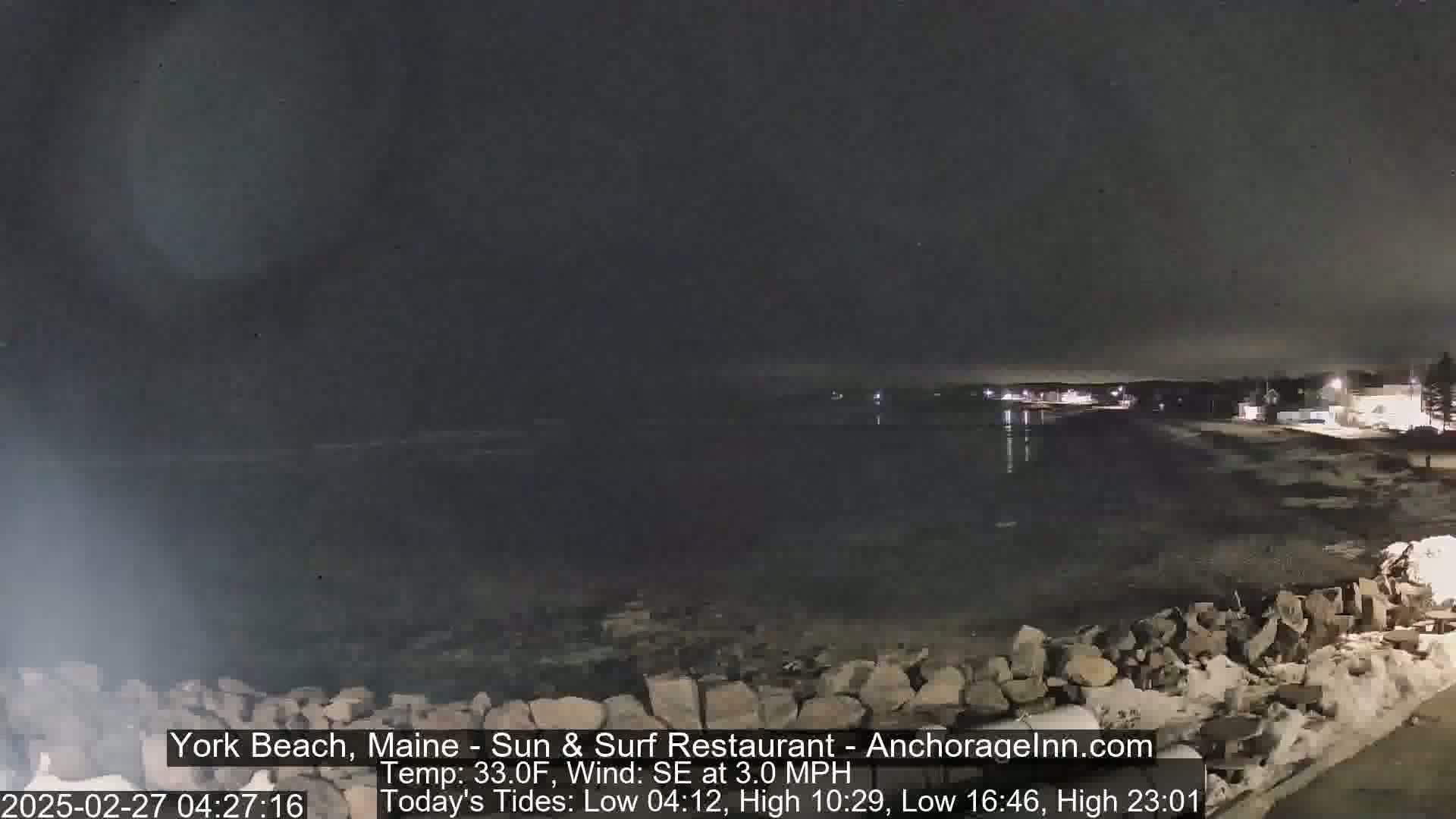 A nighttime coastal scene shows a dark body of water lapping against a rocky shoreline with a few lights visible in the distance under a dark sky.