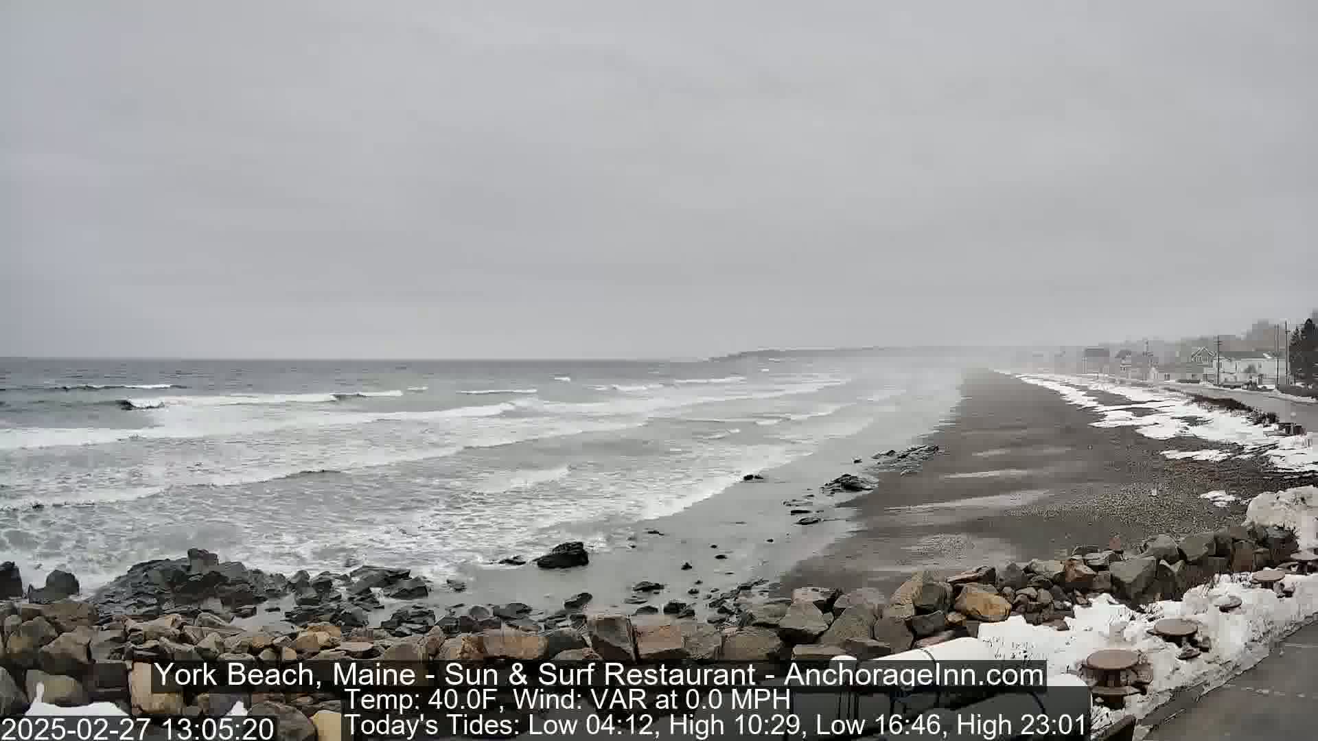 A gray, overcast day shows a beach with moderate waves, rocky shoreline, and a partially snow-covered road with buildings in the distance.