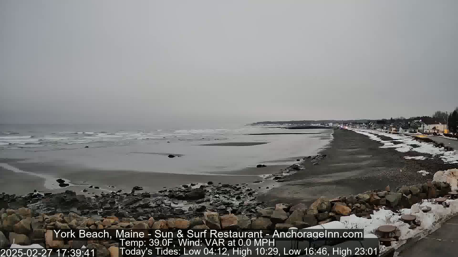 A grey, overcast day shows a dark sandy beach with small waves, a rocky shoreline, and a town visible in the distance, partially covered in snow.