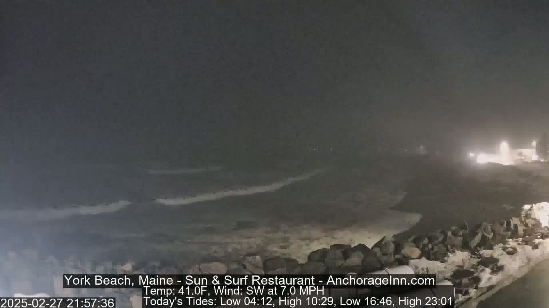 A dark, nighttime coastal scene shows a rocky shoreline with waves breaking against it under a hazy sky.