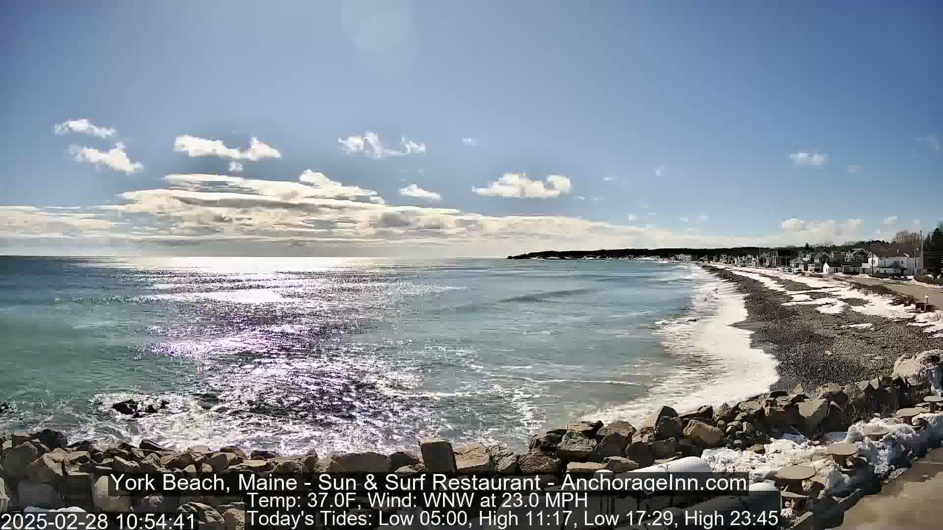 A rocky shoreline with a partially snow-covered beach meets a blue ocean with small waves under a mostly sunny sky.