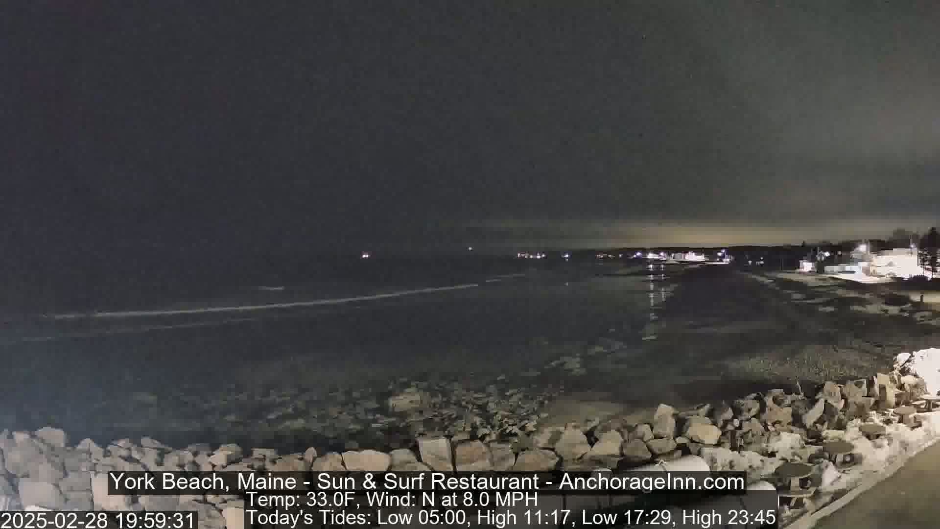 A nighttime view of a rocky shoreline and dark ocean under a mostly clear sky, with faint lights visible in the distance.