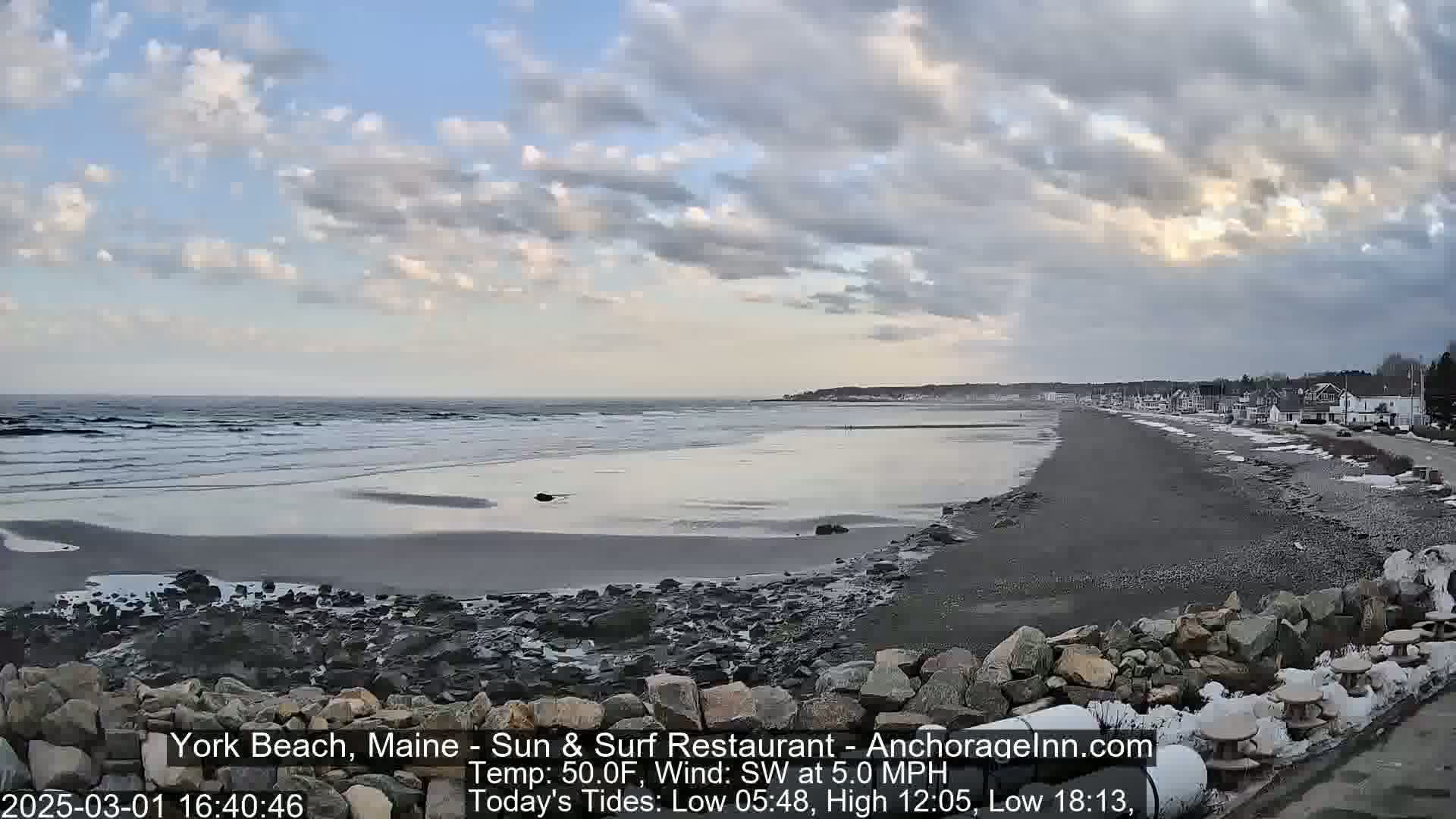 A mostly cloudy sky overlooks a long, sandy beach with gentle waves lapping the shore, backed by a line of houses and some patchy snow.