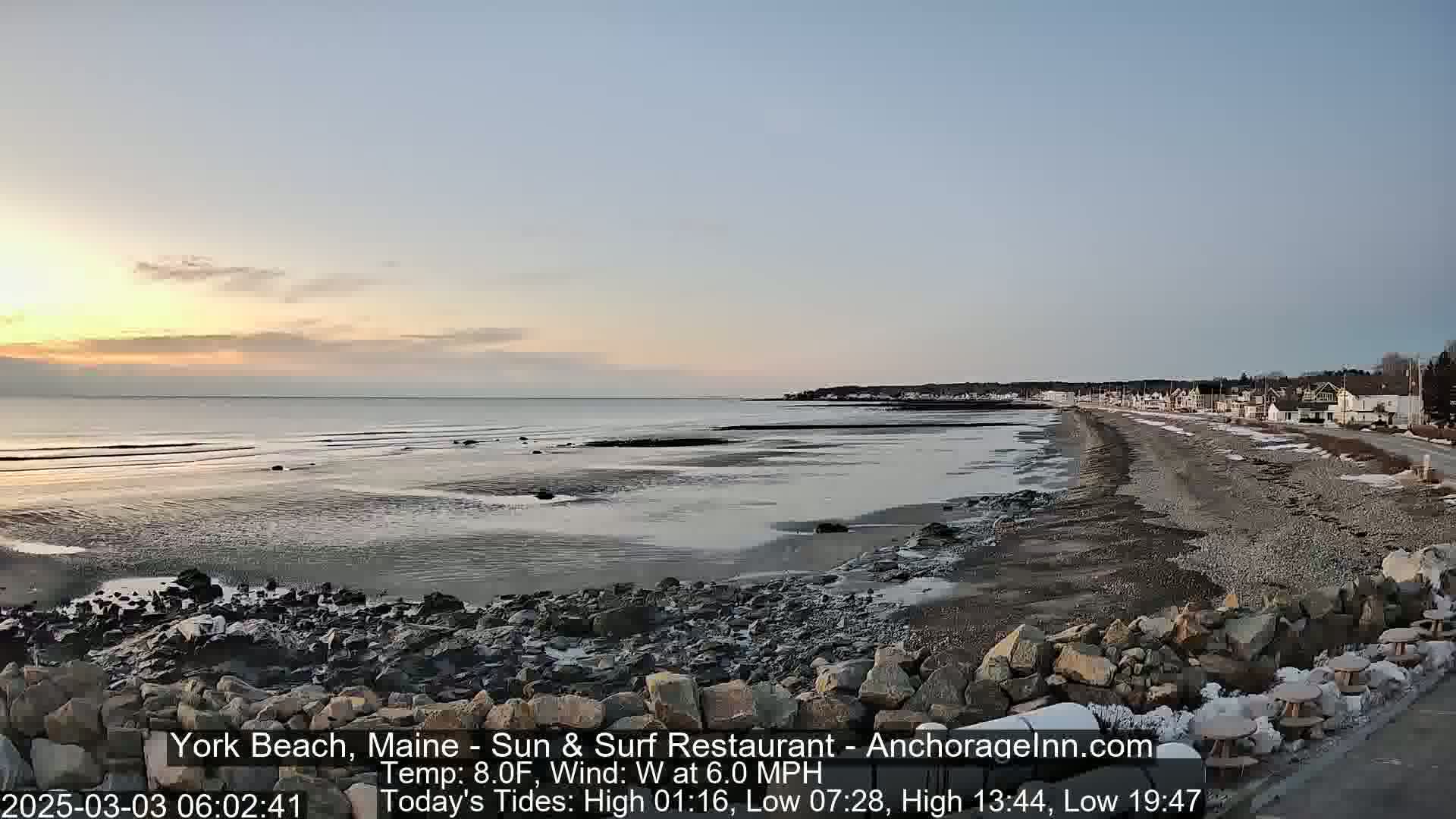 A calm, sunlit beach scene at dawn with a rocky shoreline, a calm ocean, and houses visible in the distance.