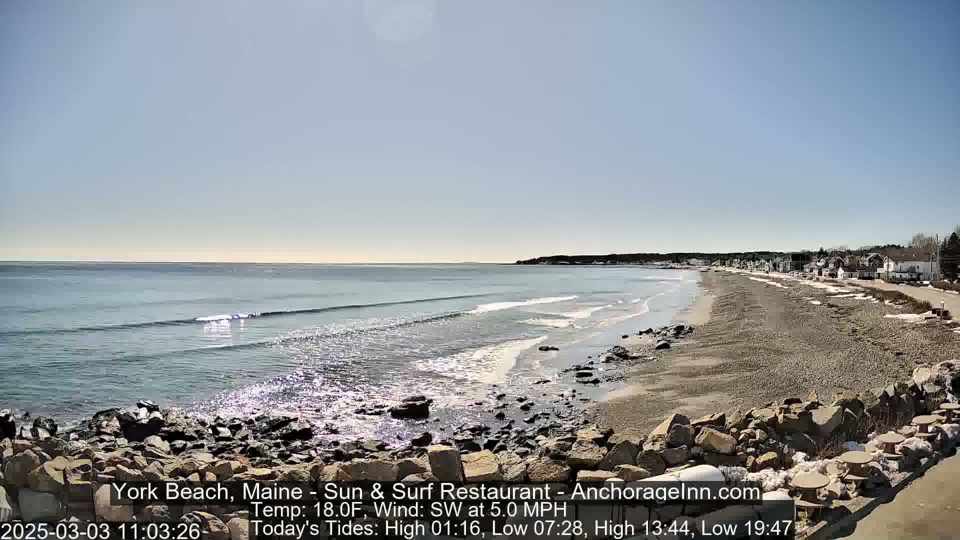 A rocky shoreline meets a sandy beach with gentle waves lapping under a clear, sunny sky.