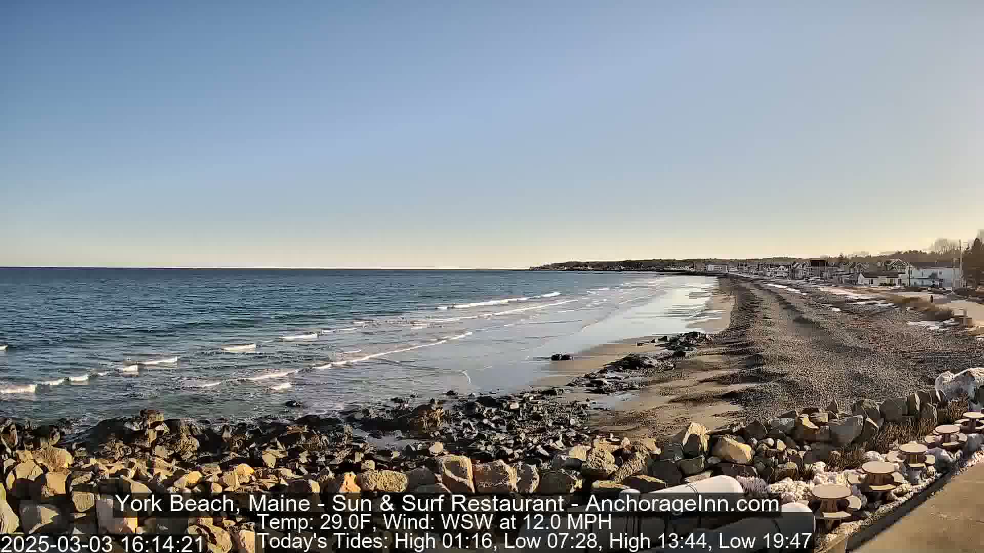 A rocky shoreline with a sandy beach stretches along a calm ocean under a clear blue sky.