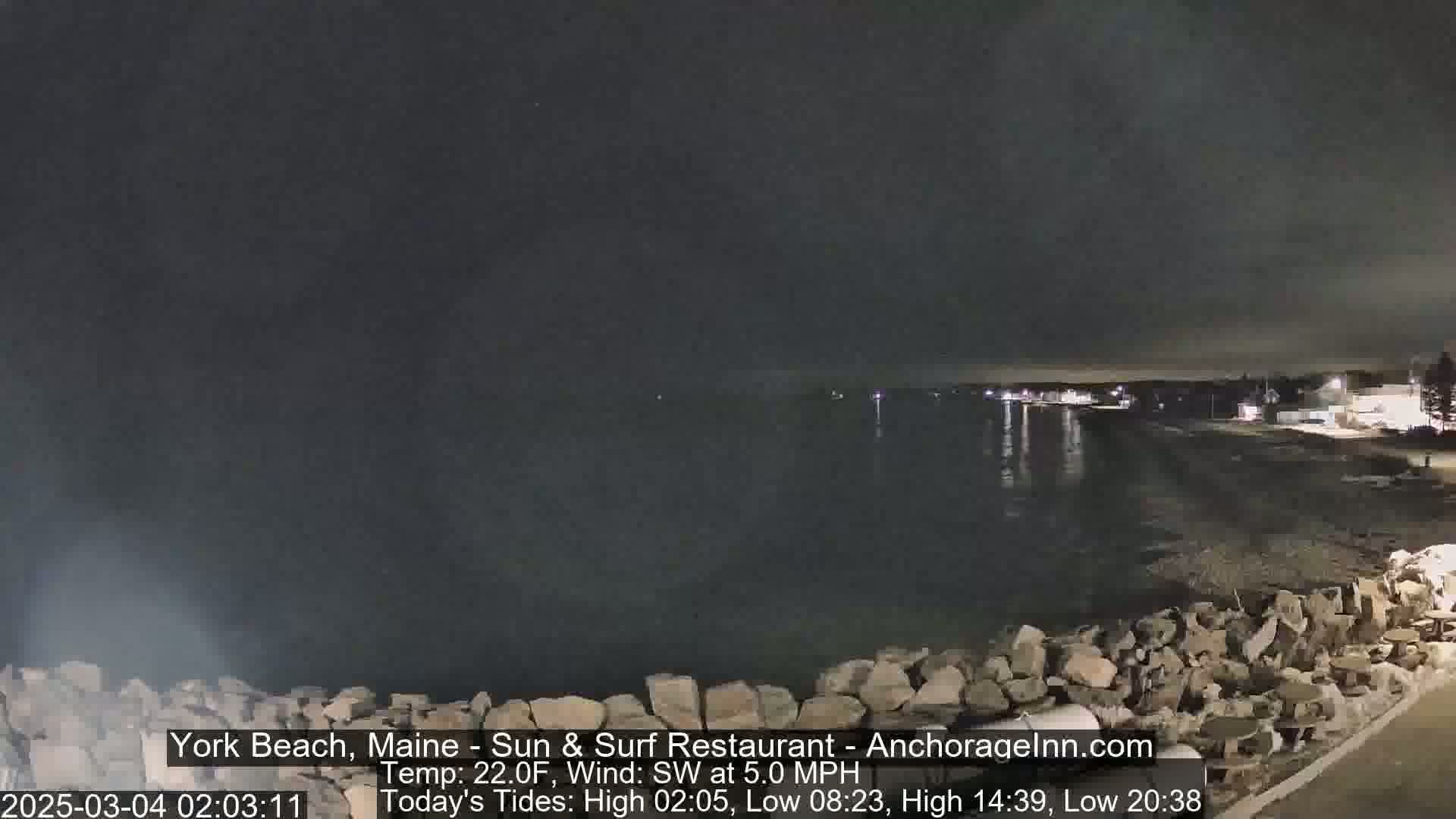 A nighttime coastal scene shows a dark body of water lapping against a rocky shore under a mostly clear sky.