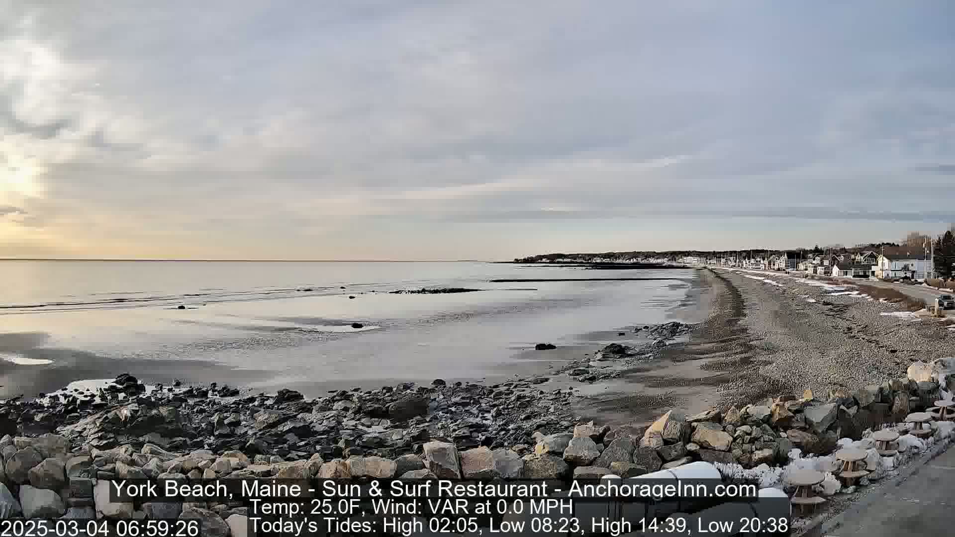 A rocky shoreline stretches along a calm, gray ocean under a mostly cloudy sky at sunrise.