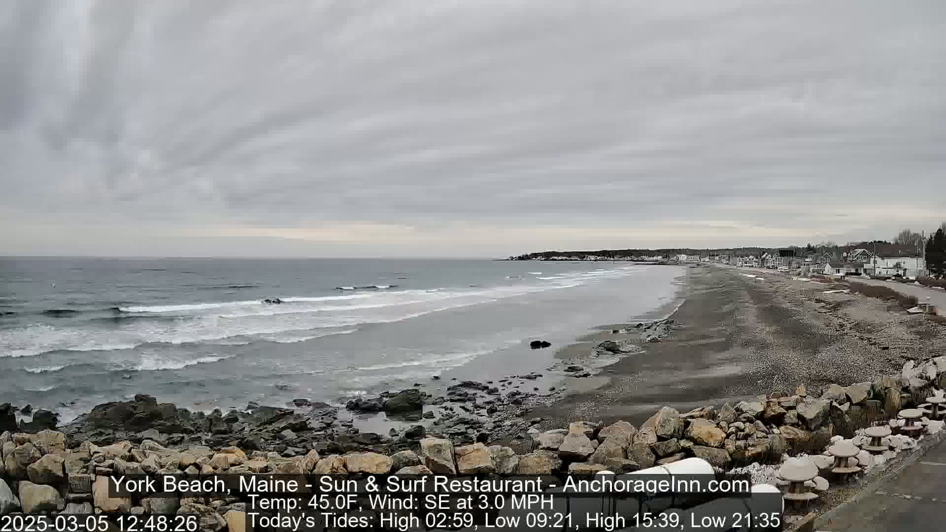 A gray, overcast day shows a beach with dark sand and rocky shoreline, gentle waves lapping the shore, and a few buildings visible in the distance.
