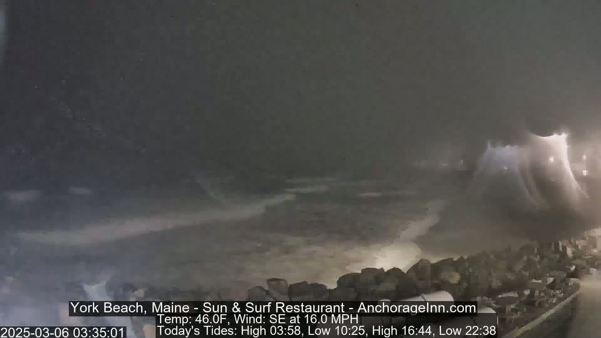 A nighttime coastal scene shows a dark, stormy ocean with large waves crashing against a rocky shoreline under a cloudy sky.