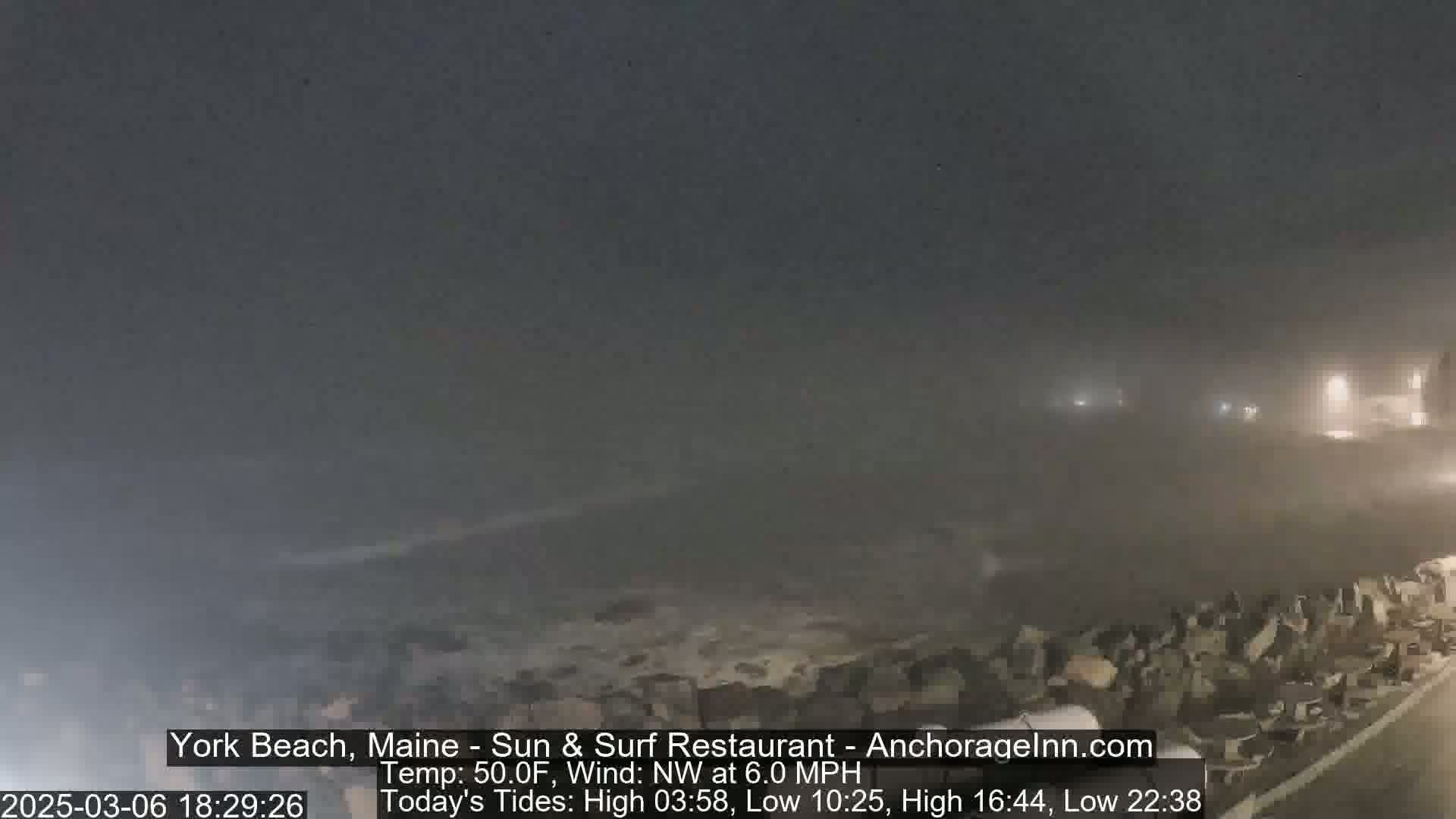 A nighttime coastal scene shrouded in fog shows a dark ocean with some waves and a rocky shoreline.