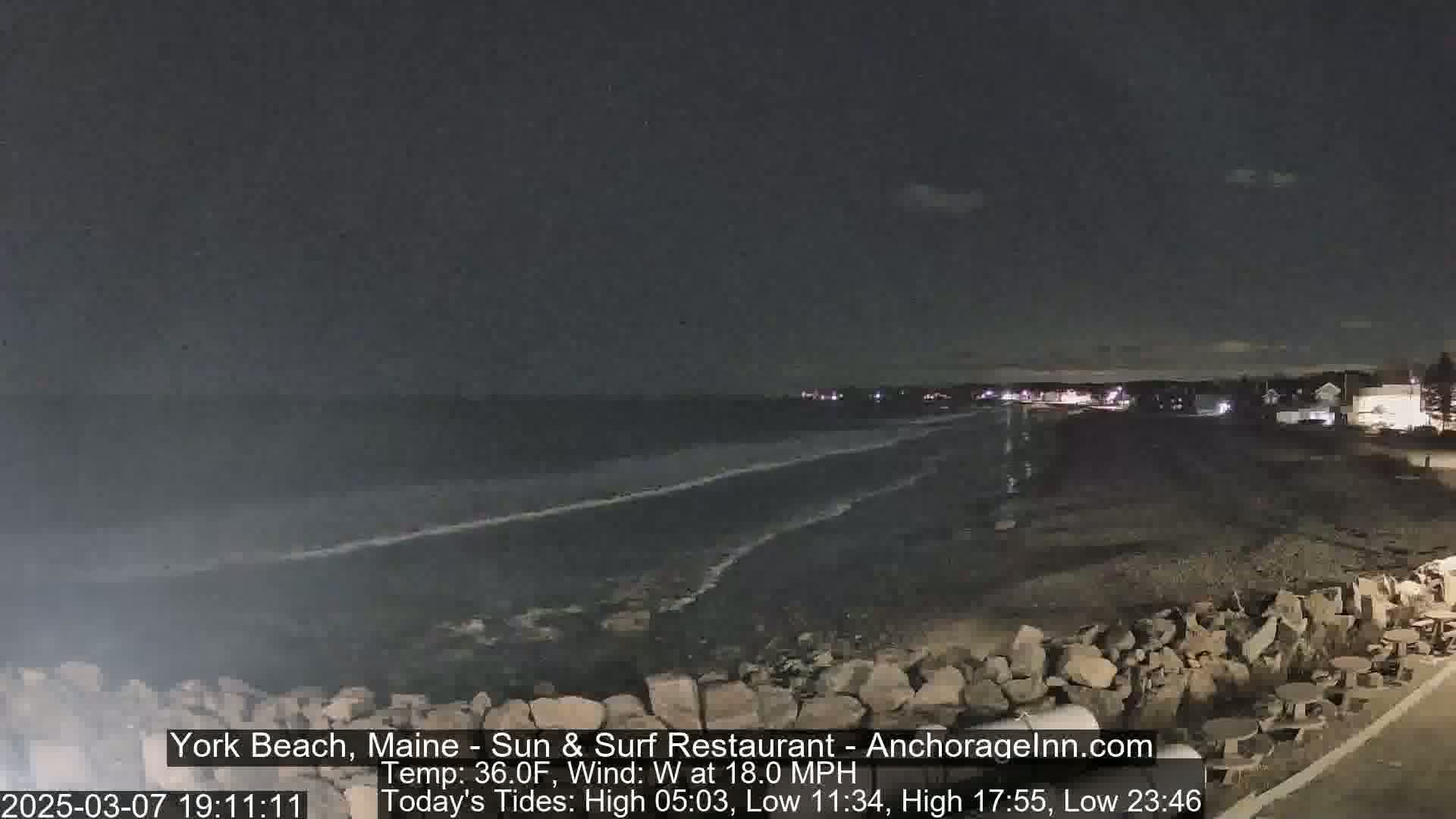 A dark nighttime beach scene shows a rocky shoreline and gently lapping waves under a mostly clear sky with a west wind at 18 mph.