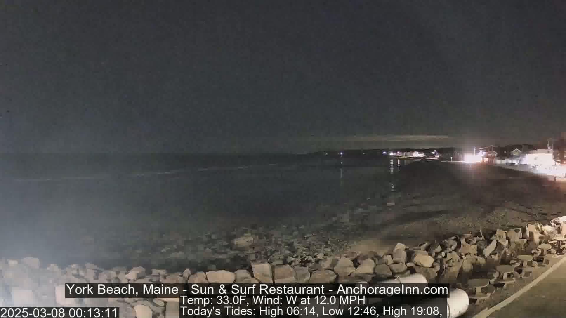 A dark nighttime scene shows a rocky shoreline and calm ocean under a clear, dark sky.
