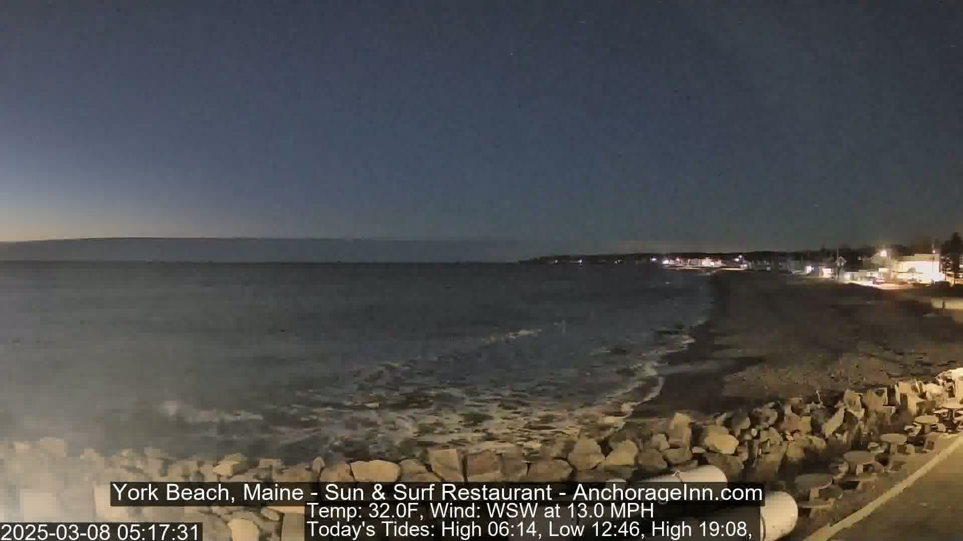 A dark, nighttime beach scene shows a rocky shoreline with gentle waves lapping the shore under a clear, dark sky.